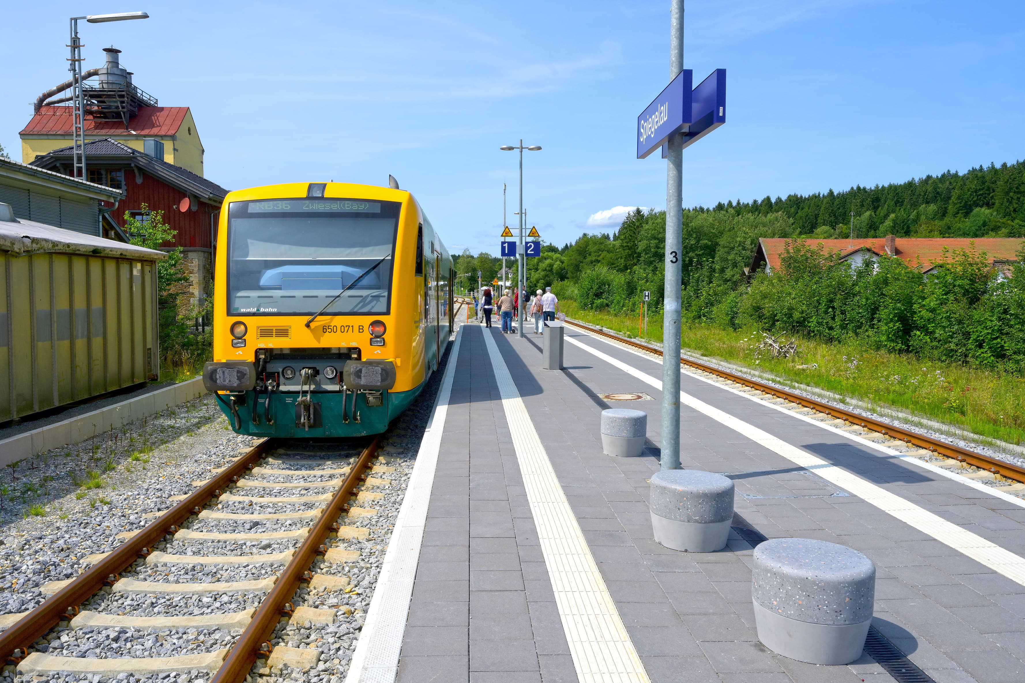 Links im Bild ein Zug. Rechts moderne Sitzhocker aus Beton auf dem neuen Bahnsteig inkl. taktilem Leitsystem. 