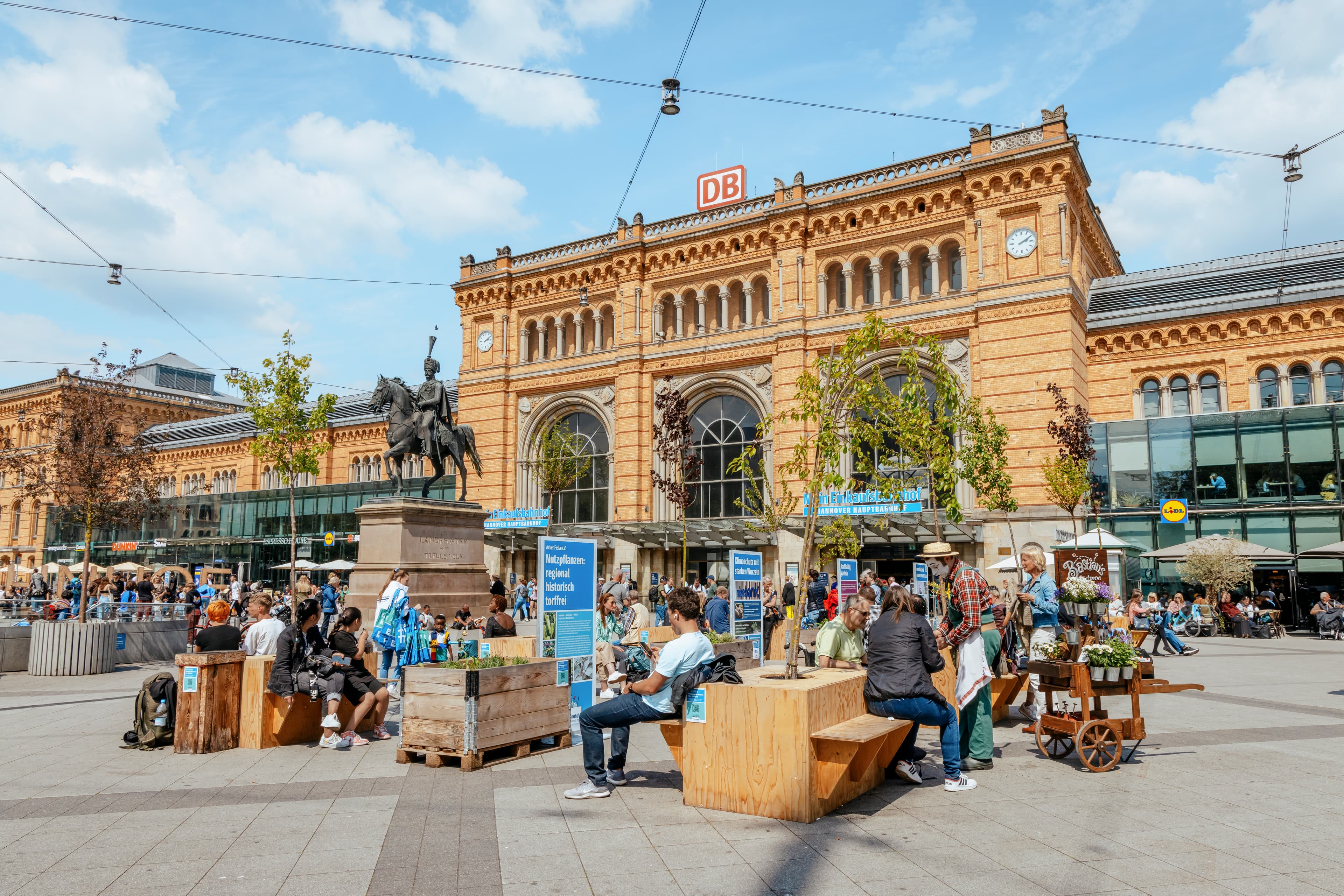 Auf dem Bahnhofsvorplatz des Hauptbahnhof Hannover sind bepflanzten Sitzmöbel, auf denen viele Menschen sitzen- Im Hintergrund sind das historische Bahnhofsgebäude und das Reiterdenkmal von Ernst-August zu sehen.