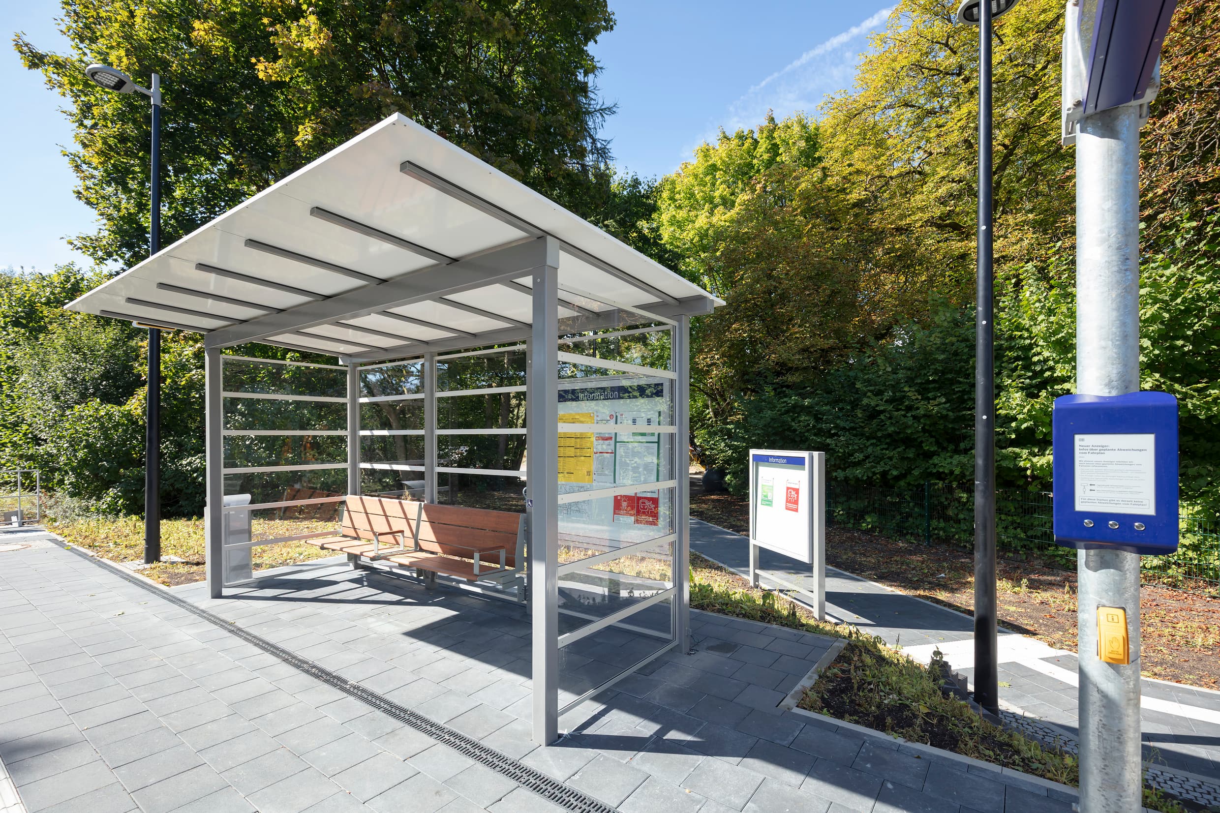 Ein transparentes Wetterschutzhäuschen mit hölzernen Bänken. Daneben die Stele vom DSA+ mit DAB und darunter der gelbe Druckknopf zur Sprachausgabe.