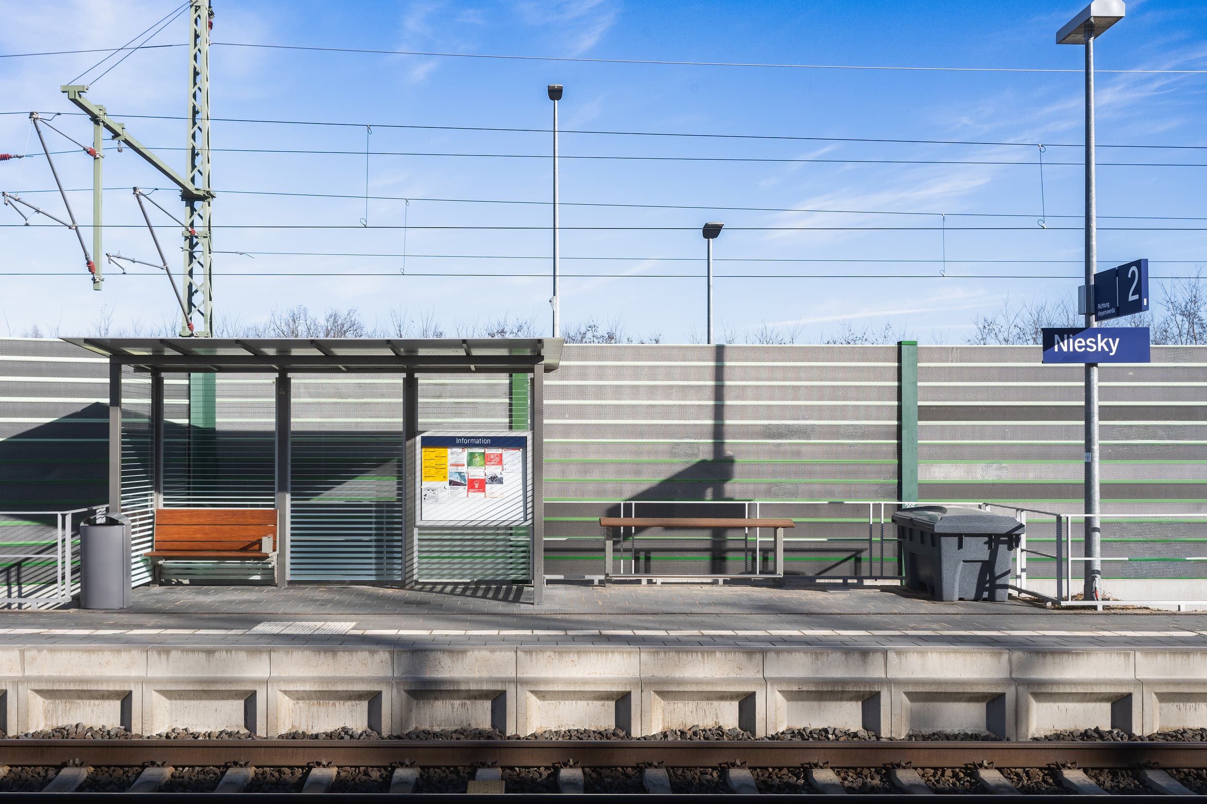 Volltransparentes Wetterschutzhaus mit neuer Holzbank und Vitrine. Daneben wurde ein zusätzlicher Anlehner aus Holz installiert. Weiter rechts steht eine Streugutkiste