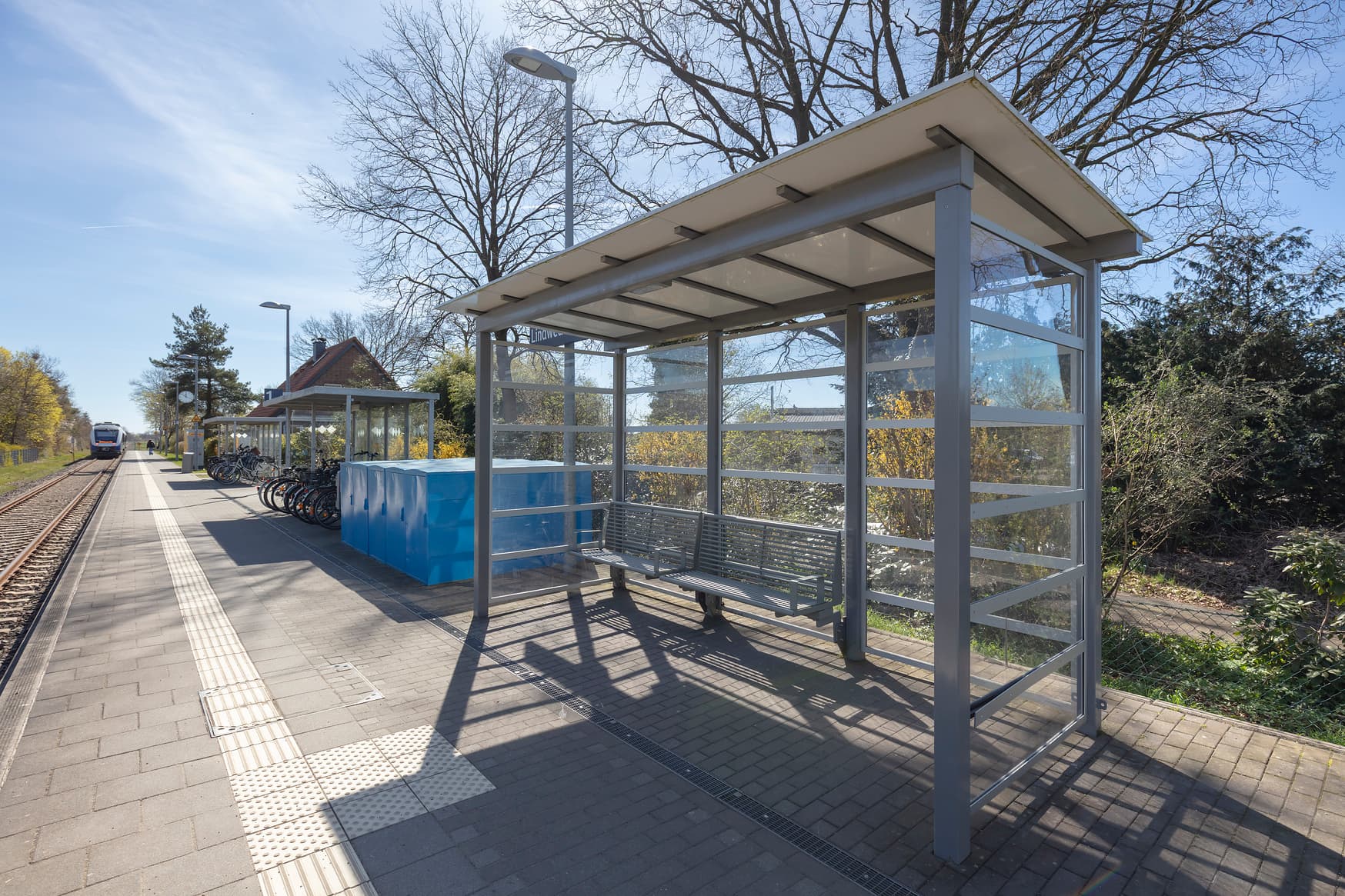Wetterschutzhäuschen mit Sitzbank an einem Bahnsteig. Im Hintergrund sind überdachte Fahrradparkplätze zu erkennen.