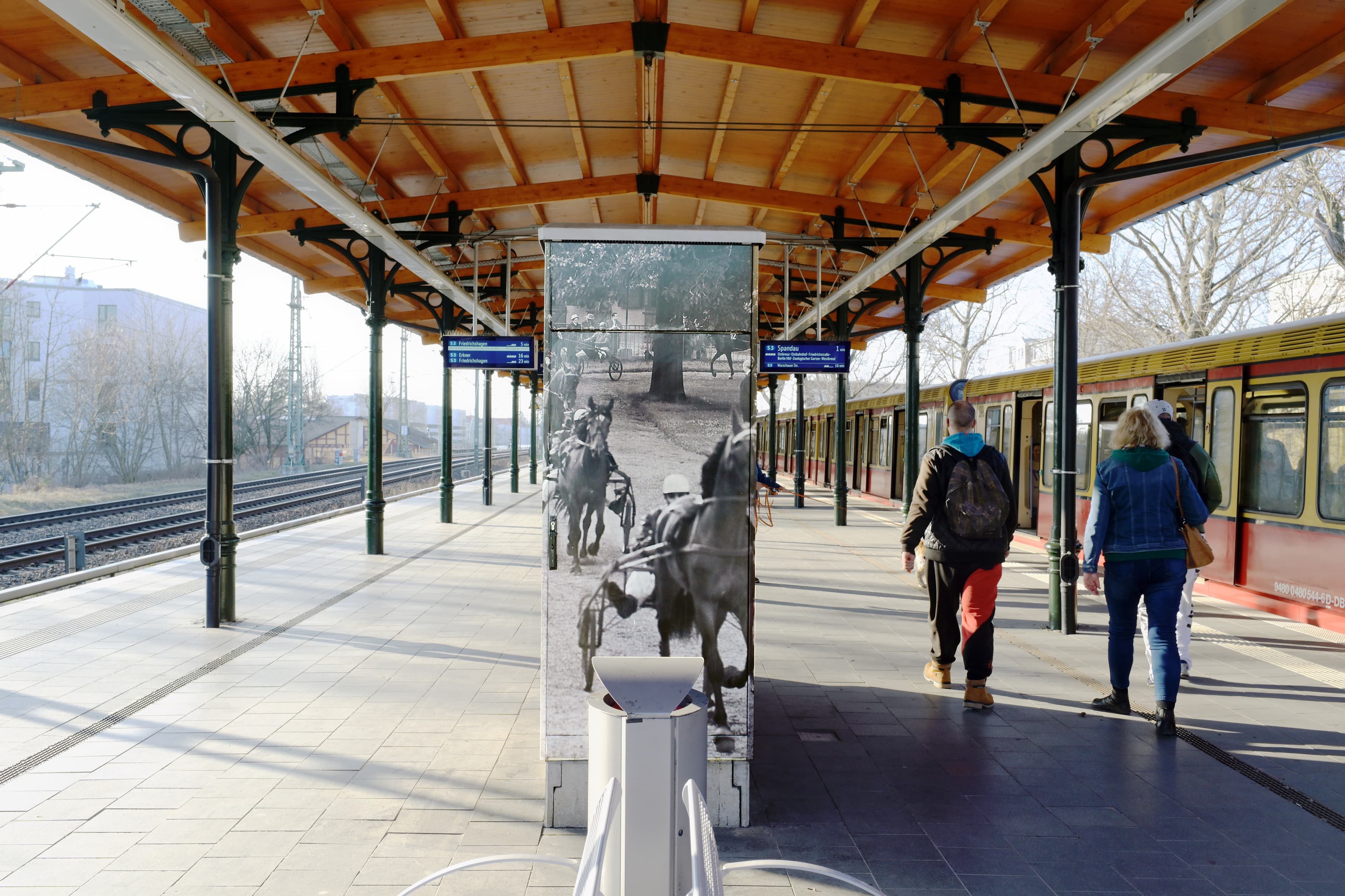 Bahnsteig mit modernen Zugzielanzeigern und Bahnsteigdach. In der Mitte befindet sich ein Snackautomat mit Motiven der Trabrennbahn.