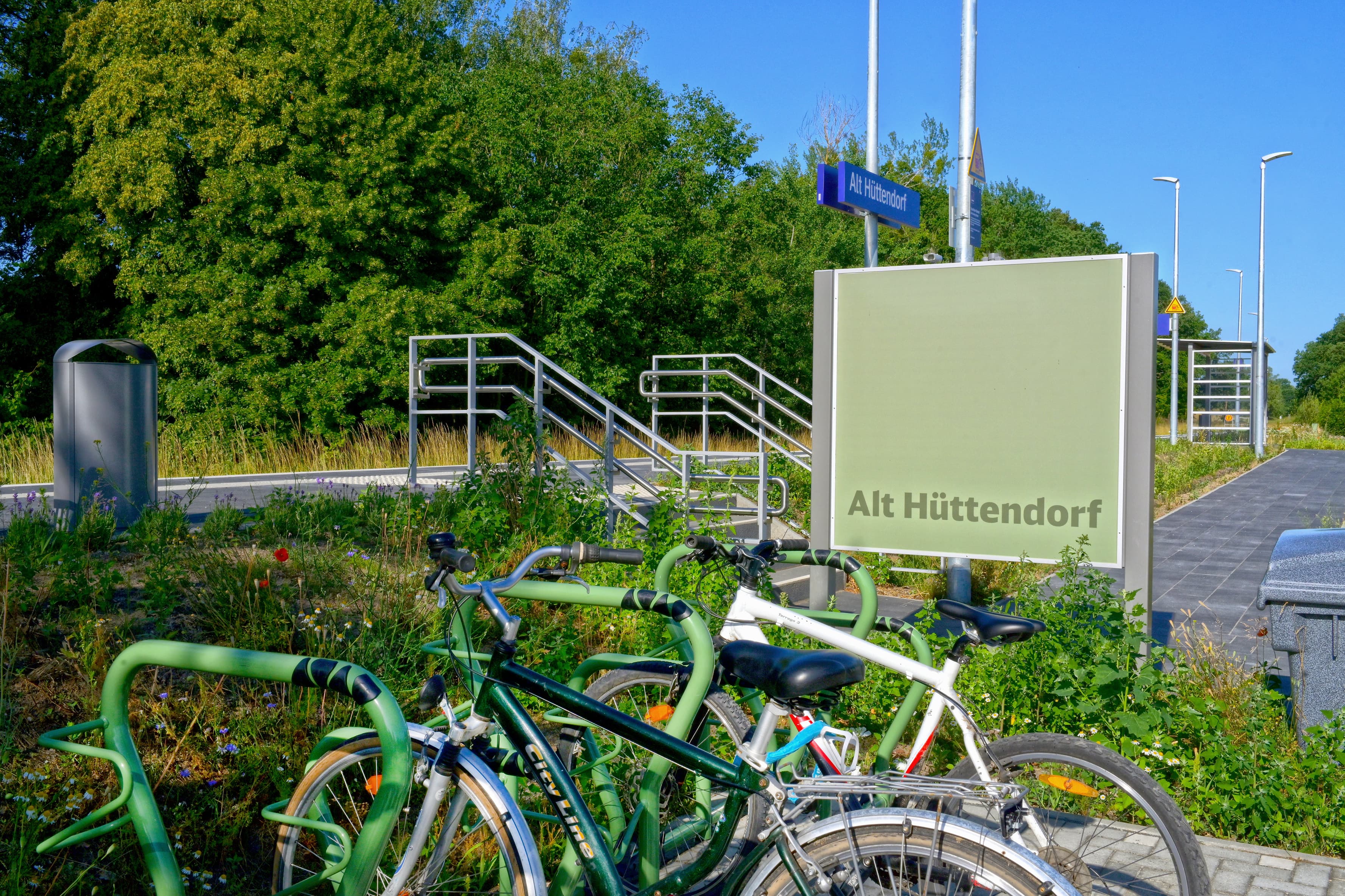 Mit dem Stationsnamen verzierte Rückseite einer Infovitrine. Davor Fahrräder an grünen Fahrradbügeln.