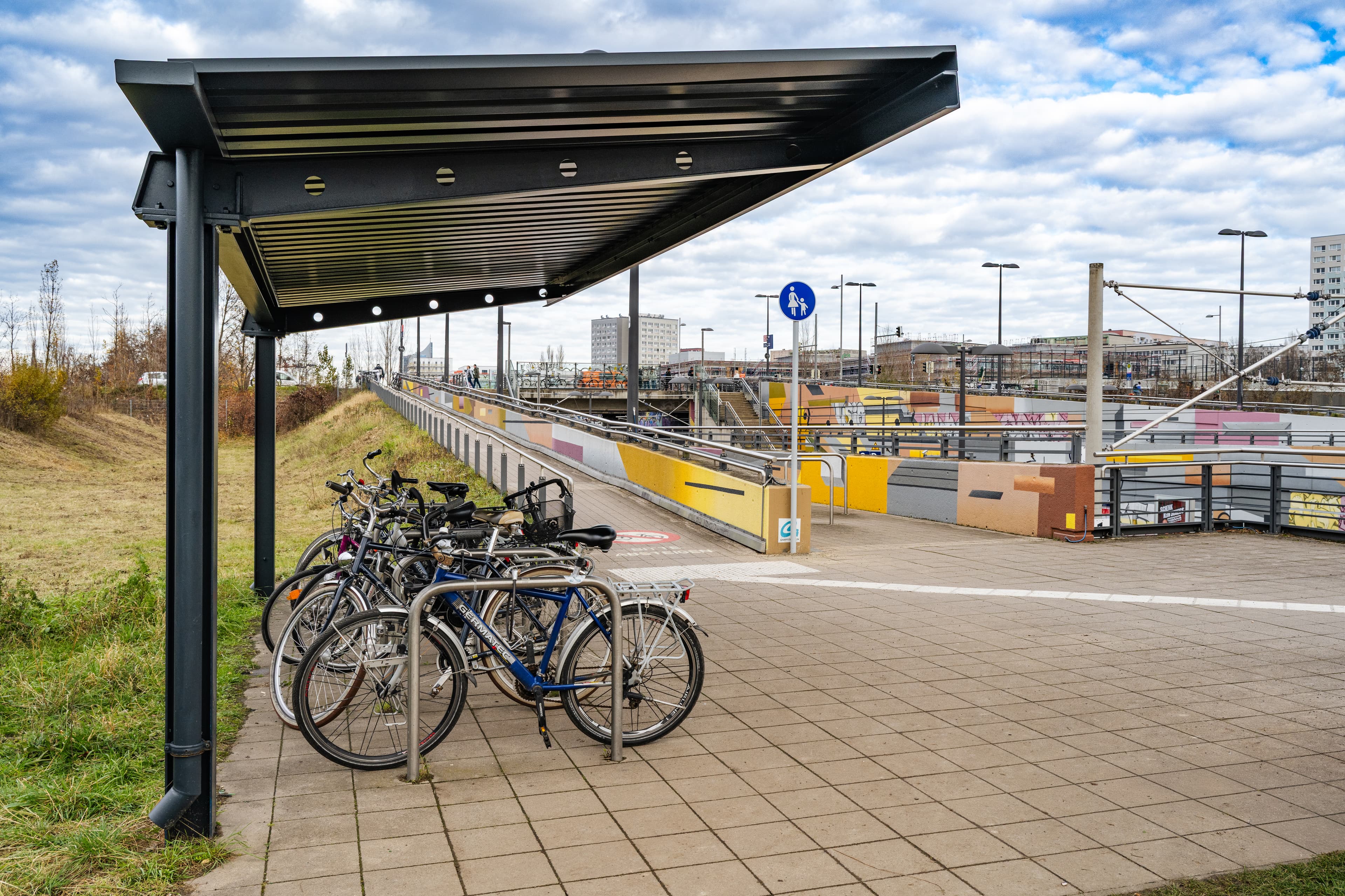 Unterhalb der Rampe am Zugang zum Bahnsteig steht die sanierte, überdachte Fahrradabstellanlage. Im Hintergrund sieht man noch die gestalteten Wände der Verkehrsstation.