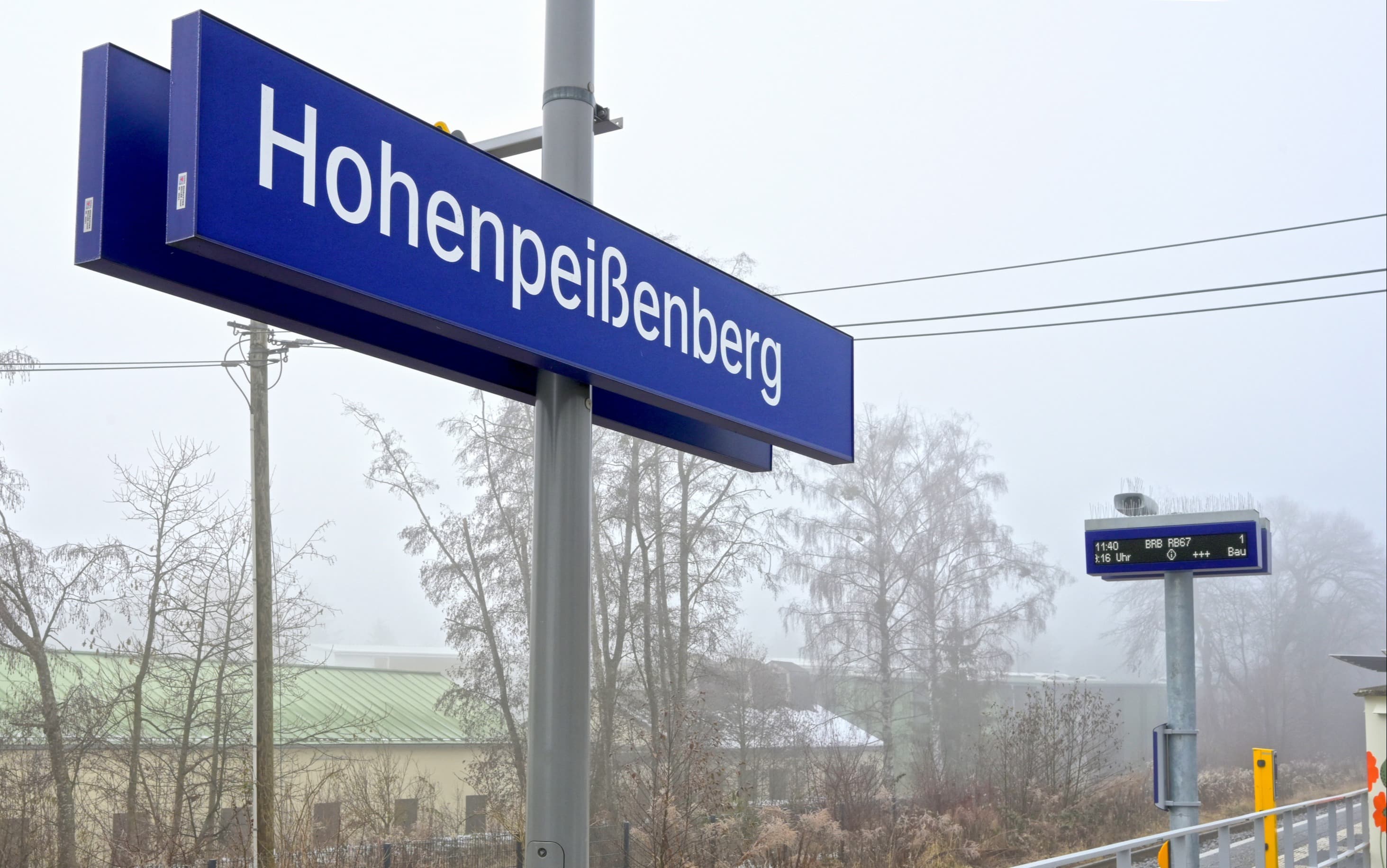 Bahnhofsschild in blau, rechts davon im Nebel immer noch erkennbar der dynamische Scriftenanzeiger.