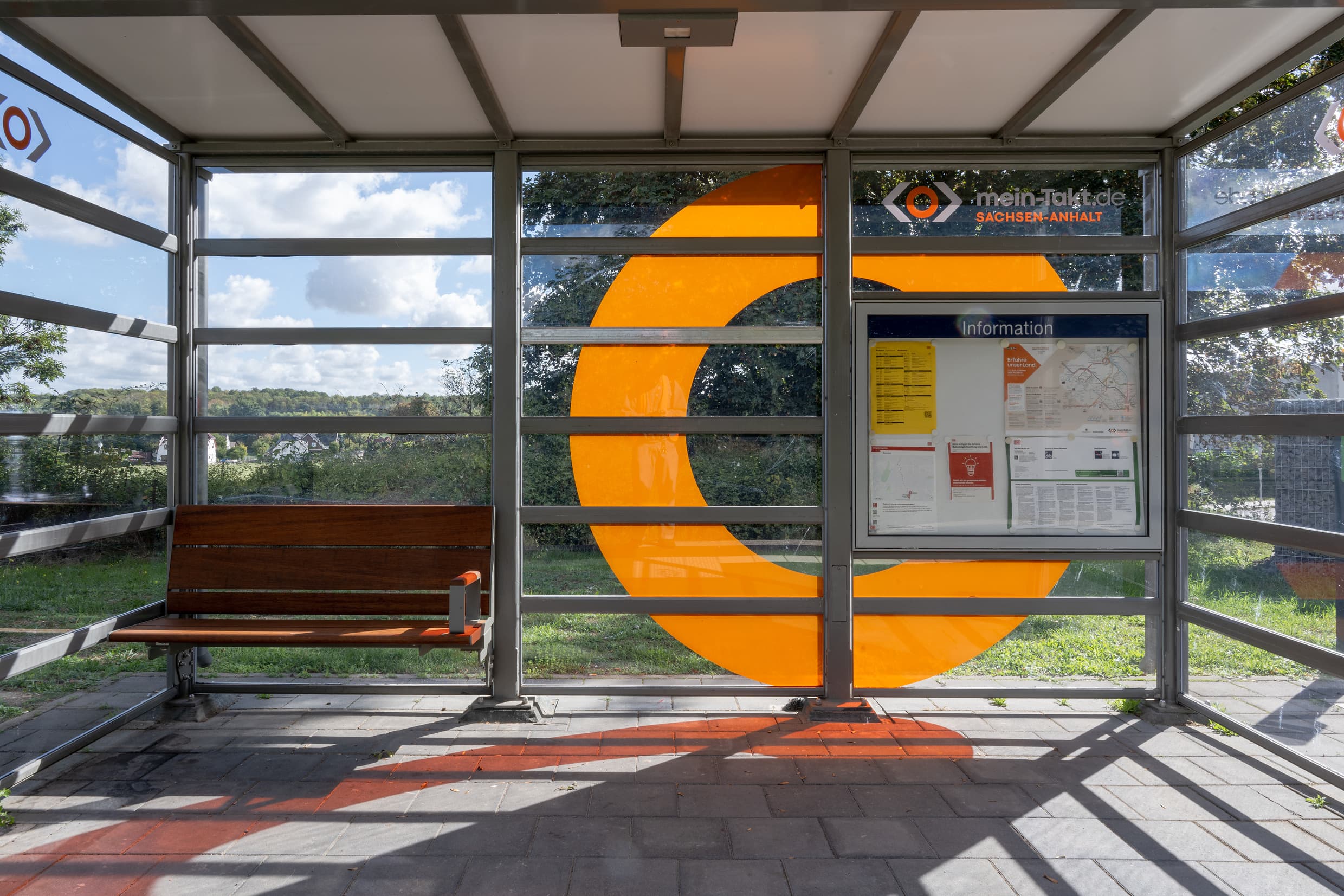Detailfoto vom Wetterschutzhaus mit inzwischen voll transparenten Schreiben inkl. eines orange farbigen Kreises und Logos des Aufgabenträgers. Außerdem sieht man die neu installierte Bambusbank und die Vitrine mit den Fahrplanaushängen.