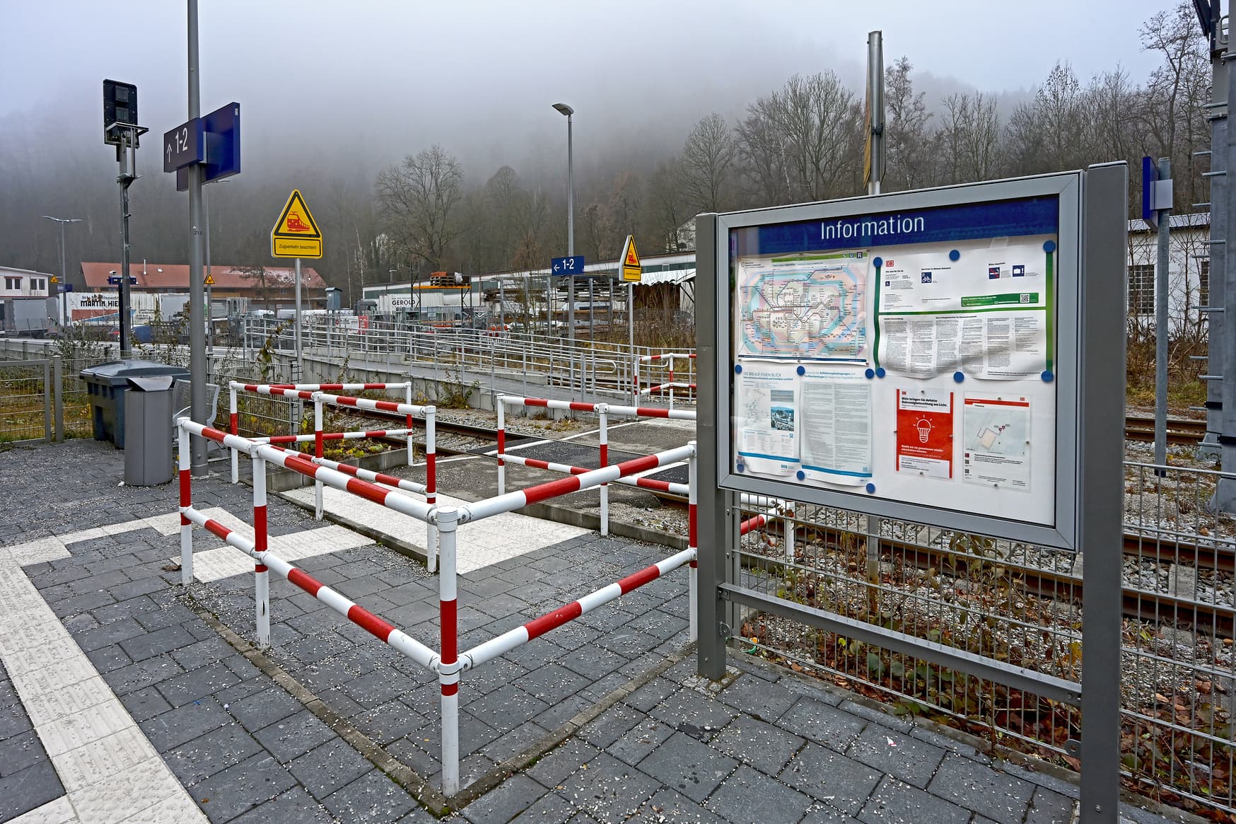 Der Bahnsteigzugang mit rot-weißem Sicherheitszaun. Daneben eine Vitrine mit aktuellen Aushängen und Informationen zum Bahnhof.