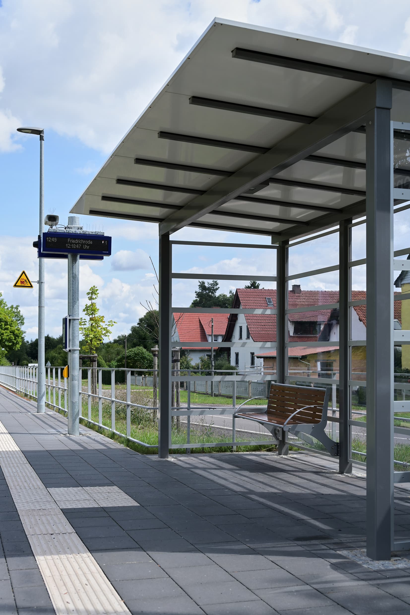 Zu sehen ist das volltransparente Wetterschutzhaus mit einer Holzbank. Im Hintergrund erkennt man den zweizeiligen digitalen Schriftanzeiger für die Reisendeninformation.