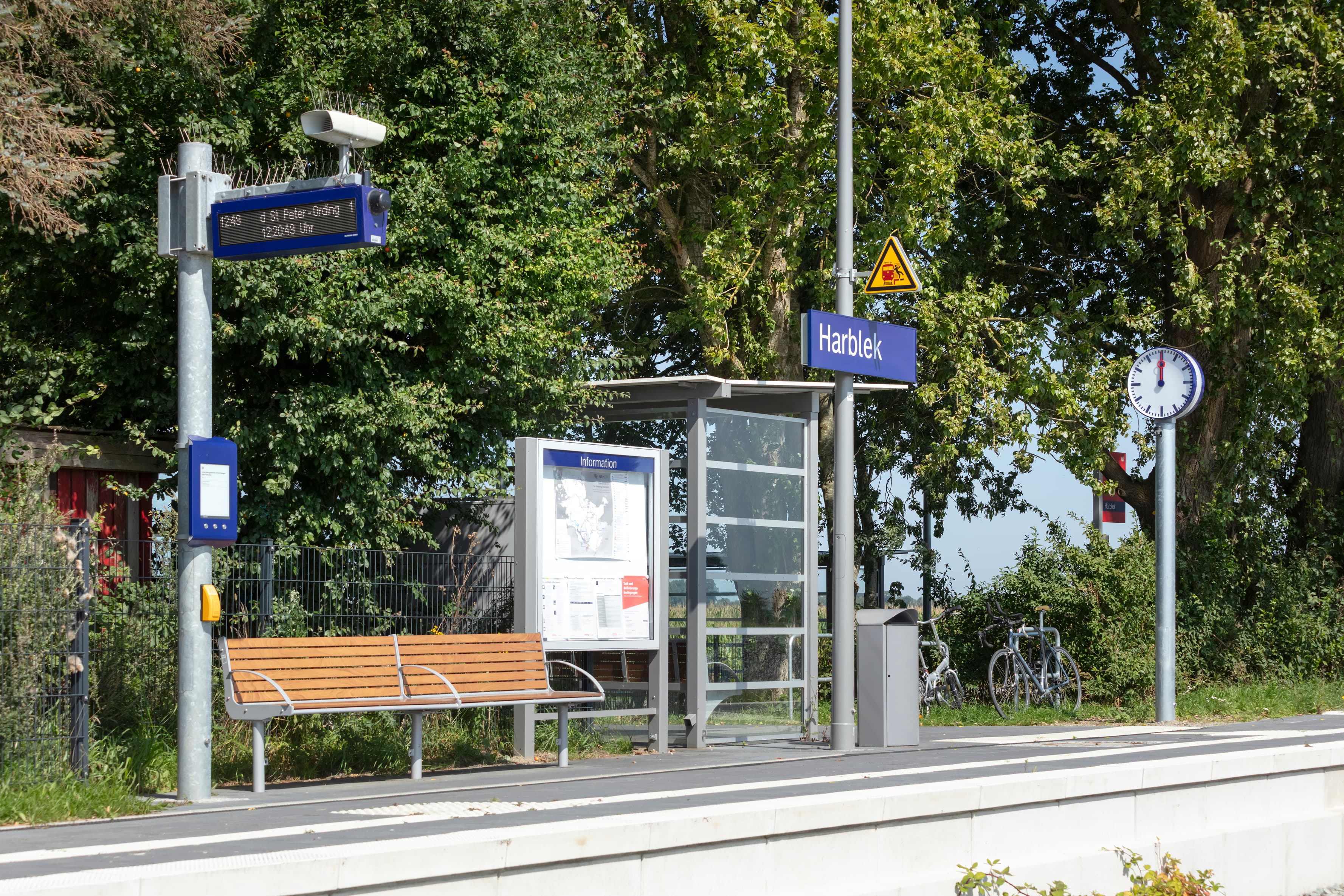 Neu gestalteter Bahnsteig mit Wetterschutz, Bänken und einem Zugzielanzeiger. Linker Hand der Blick auf Felder.