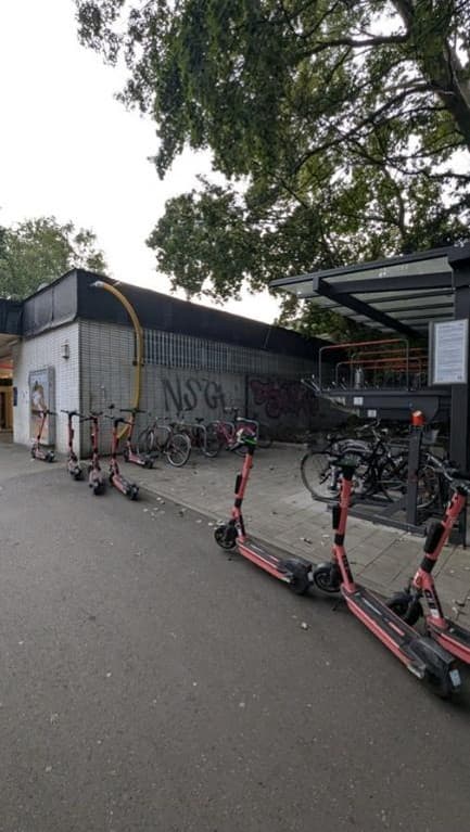 Die Seite des Empfangsgebäudes mit grauen Fliese. Davor Fahrradbügel und eine Fahrradabstellanlage. Überall stehen E-Scooter herum.