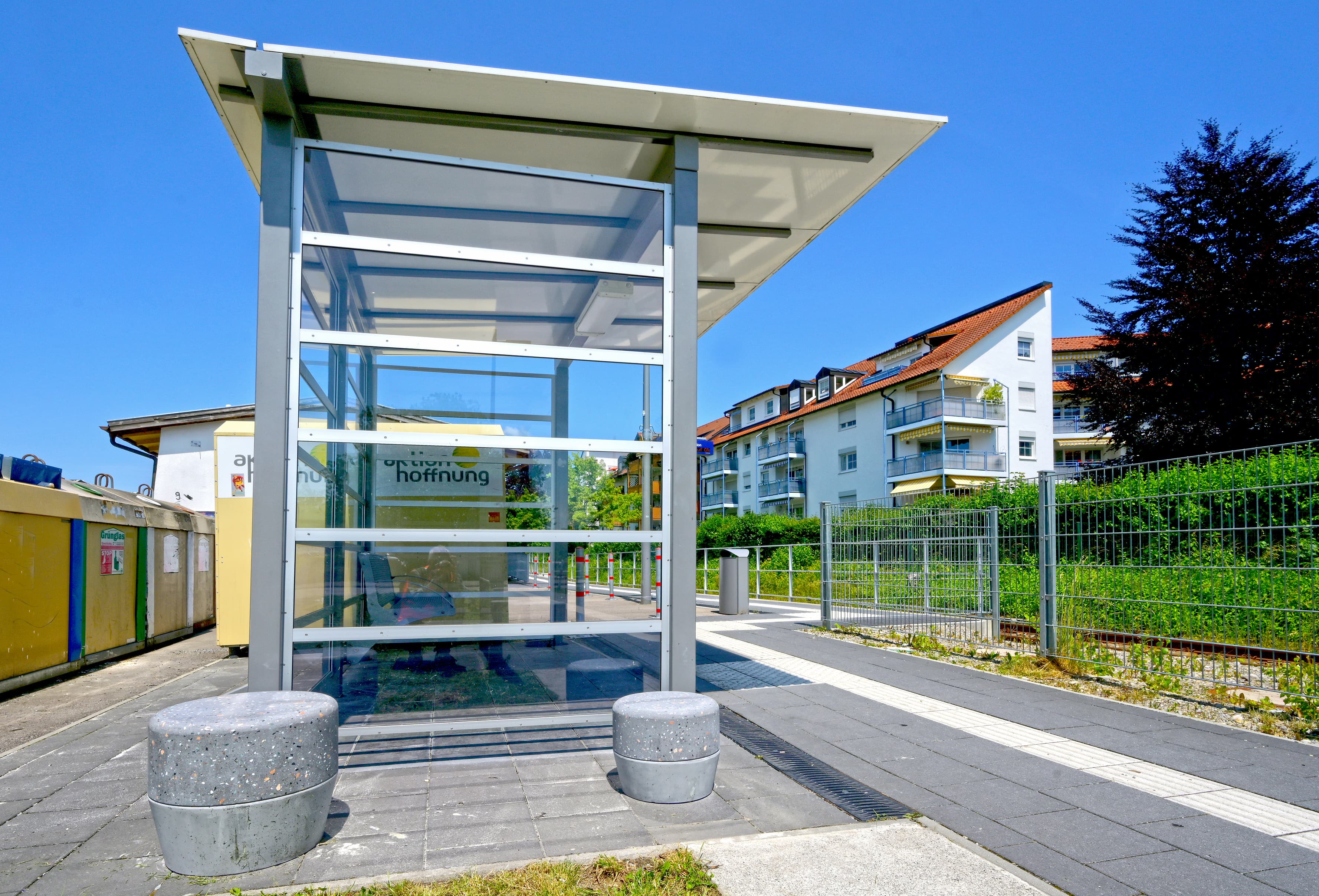 Zwei Hocker aus Beton. Dahinter ein Wetterschutzhaus mit transparenter Rahmung. Davor der Bahnsteig.
