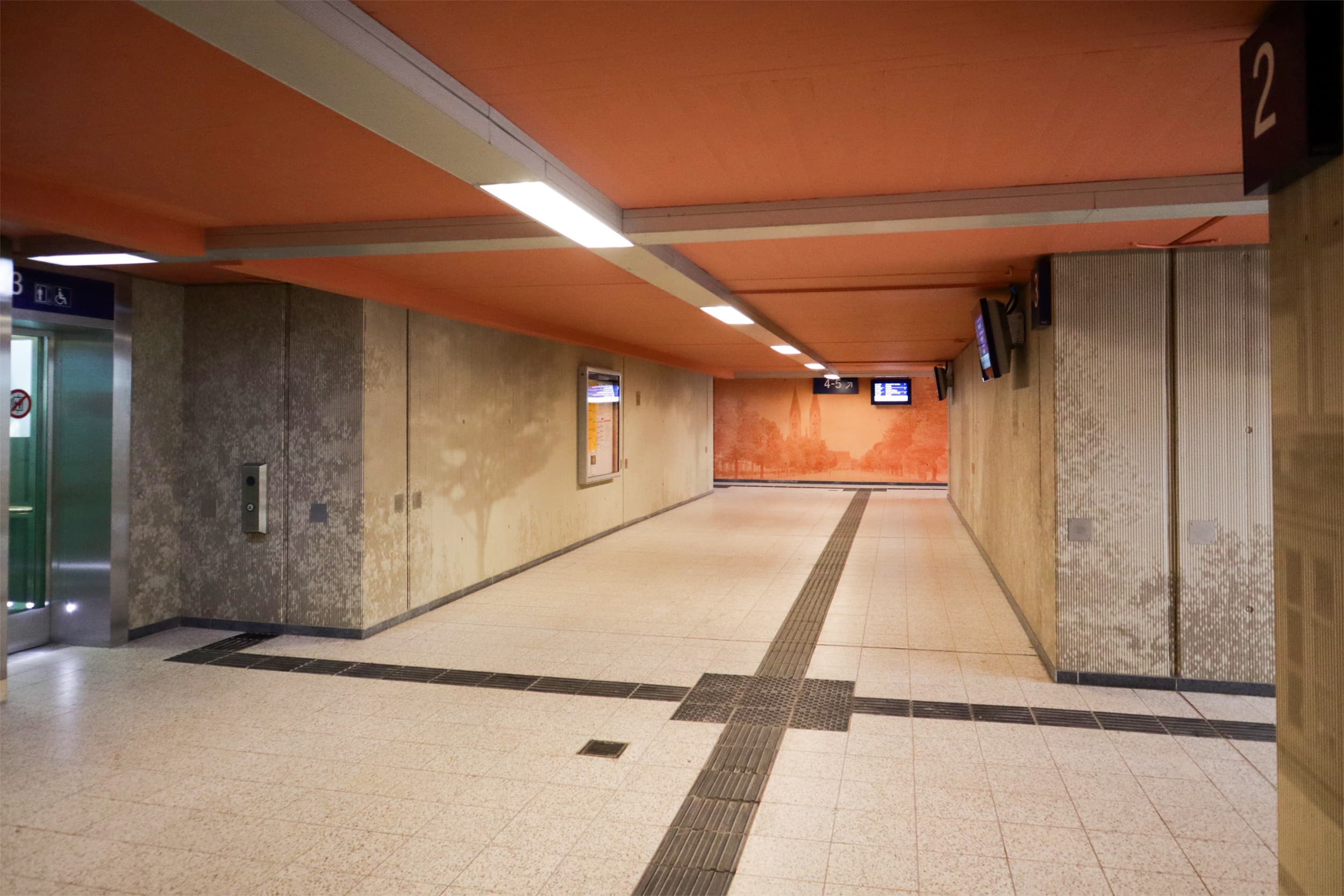 Neu gestaltete Personenunterführung mit grafischen Elementen, die die Architektur der Stadt aufnehmen, heller Beleuchtung, neuen Monitoren an jedem Aufgang und einer orangenen Deckenfarbe.