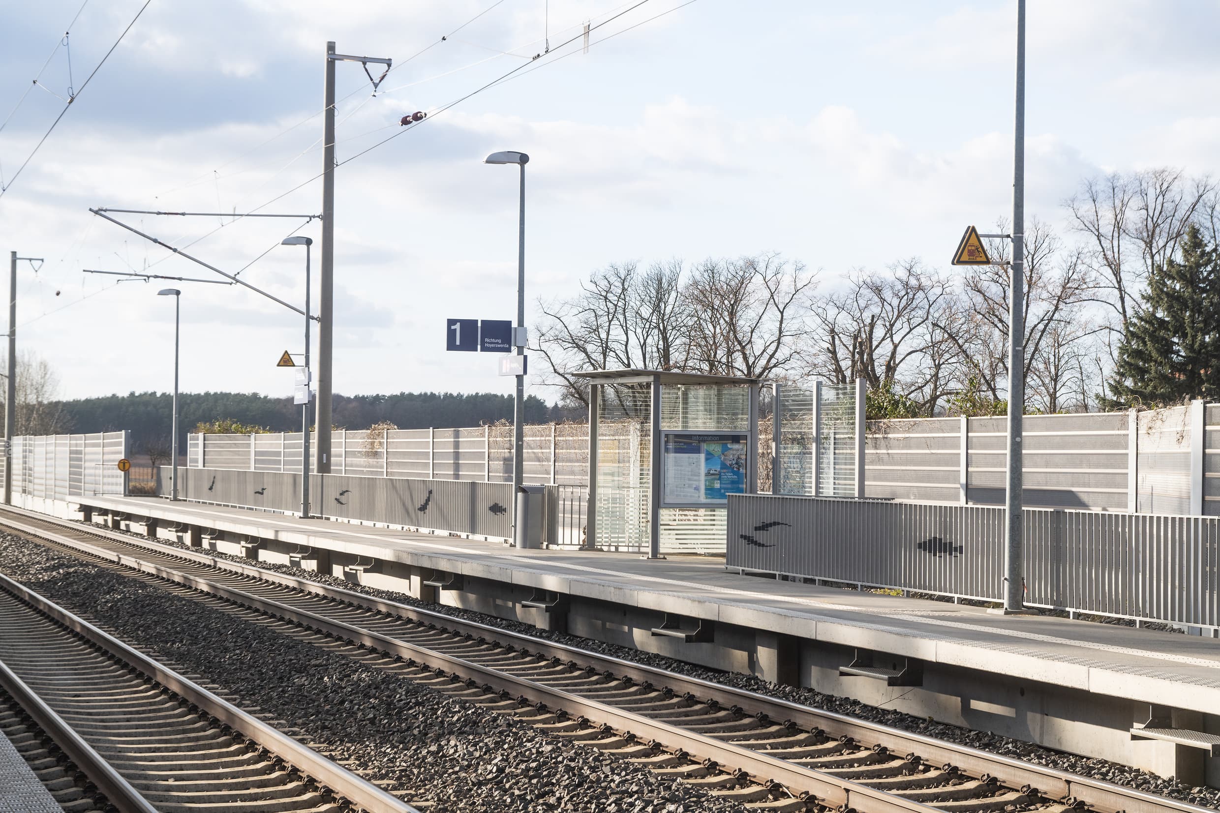 Blick auf den gesamten Bahnsteig mit Wetterschutzhäuschen. Der Bahnsteig wird durch ein graues Geländer abgegrenzt, welches mit Fischsilhouetten gestaltet wurde.