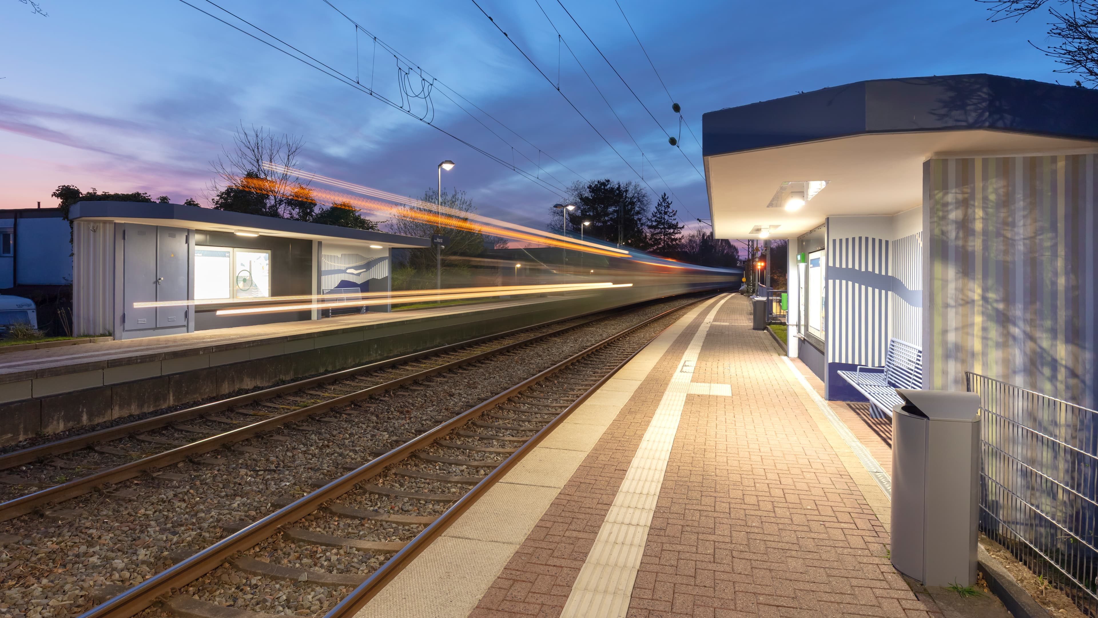 Blick auf zwei Bahngleise. Rechts und links davon je ein Bahnsteig. Ein vorbeifahrender Zug ist zu erkennen.