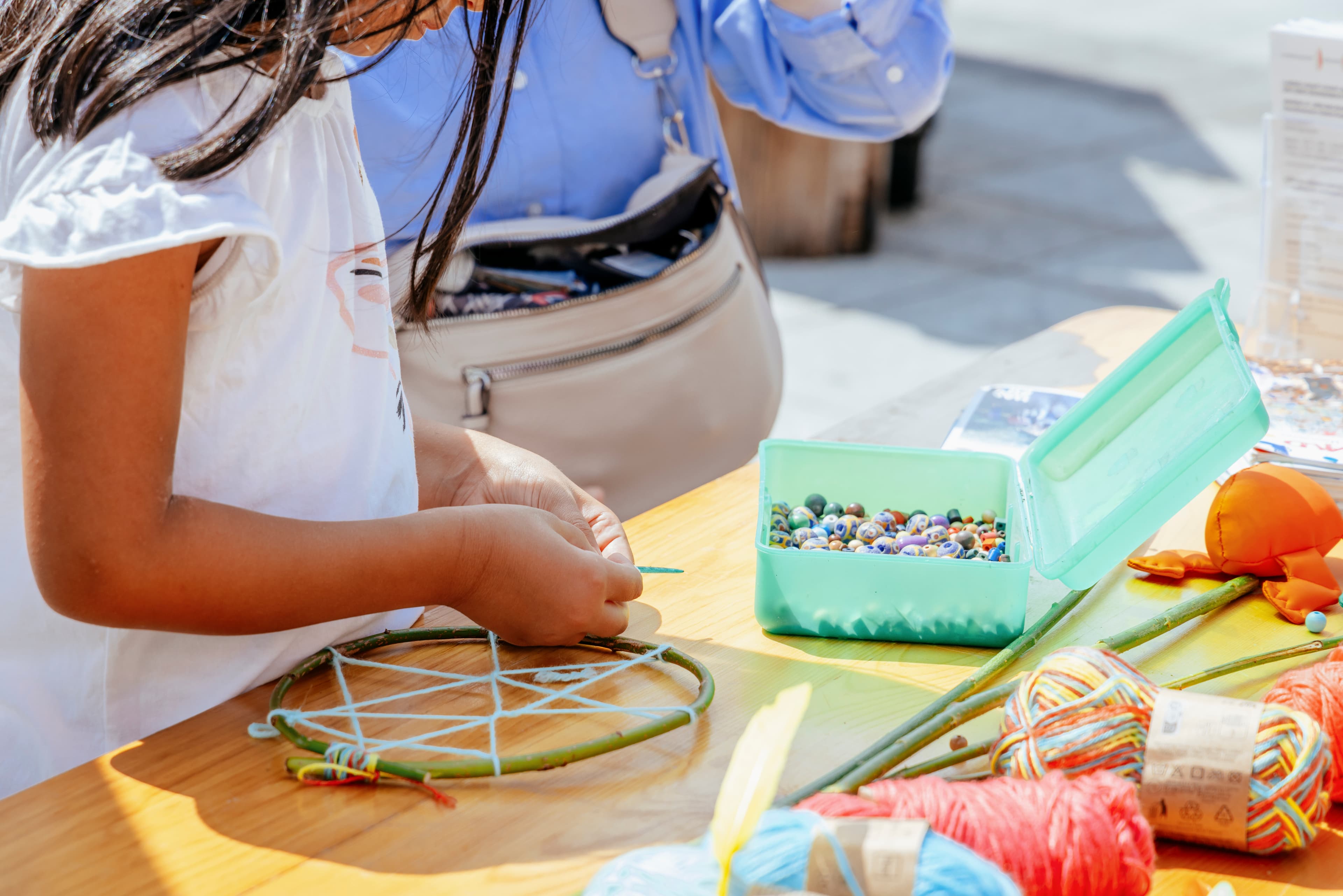 Kinderhände basteln an einem Tisch mit natürlichen Materialien wie Zweigen, Wolle und Steinen eine Art Traumfänger. Im Hintergrund ist die geöffnete Tasche eines erwachsenen Menschen zu sehen.