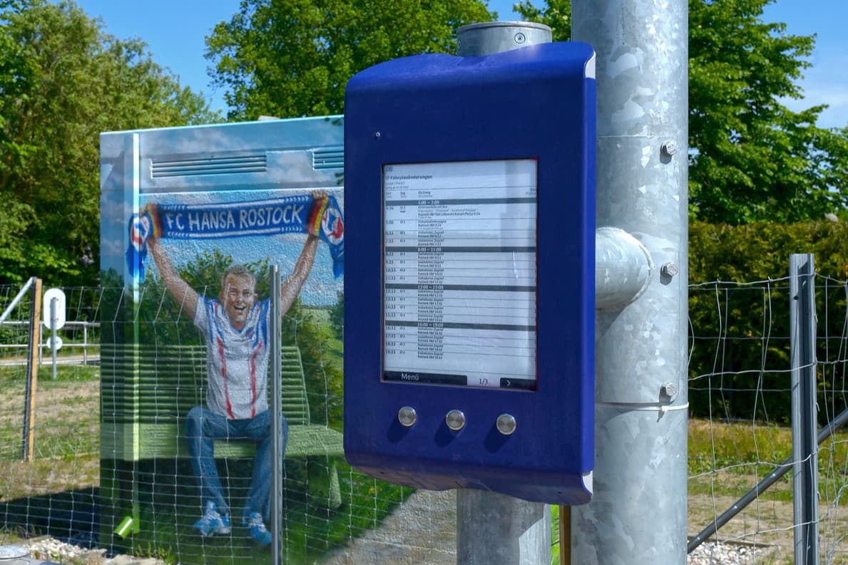 Blick auf eine blaue Anzeige am Bahnhof, im Hintergrund ein Häuschen auf dem ein Hansa Rostock Fan gemalt ist.