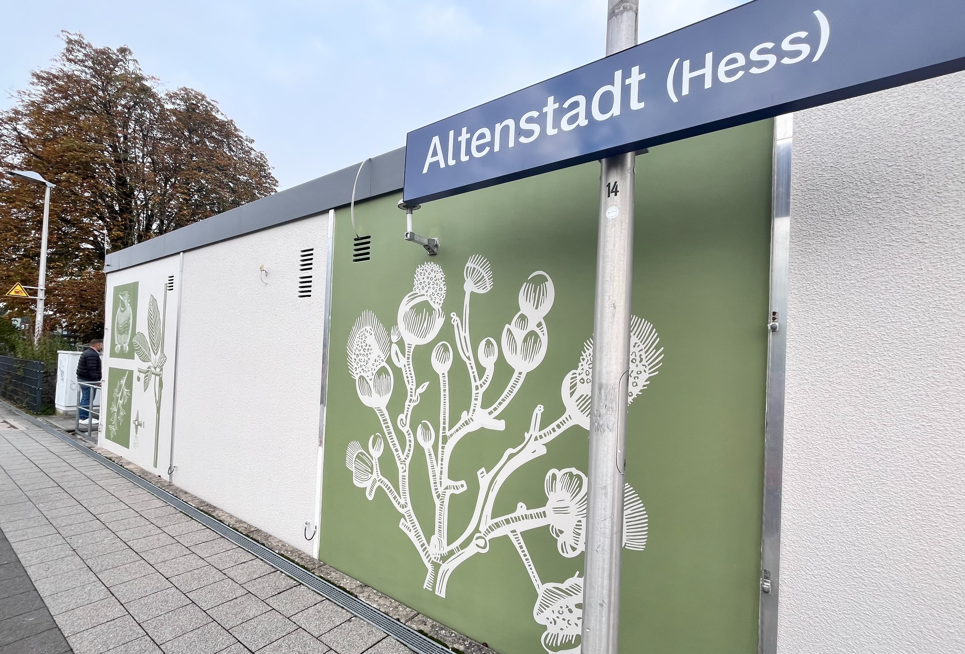 Bahnhofsschild von Altstadt( Hess) vor einem grün gestalteten Häuschen mit weißen, floralem Detail.