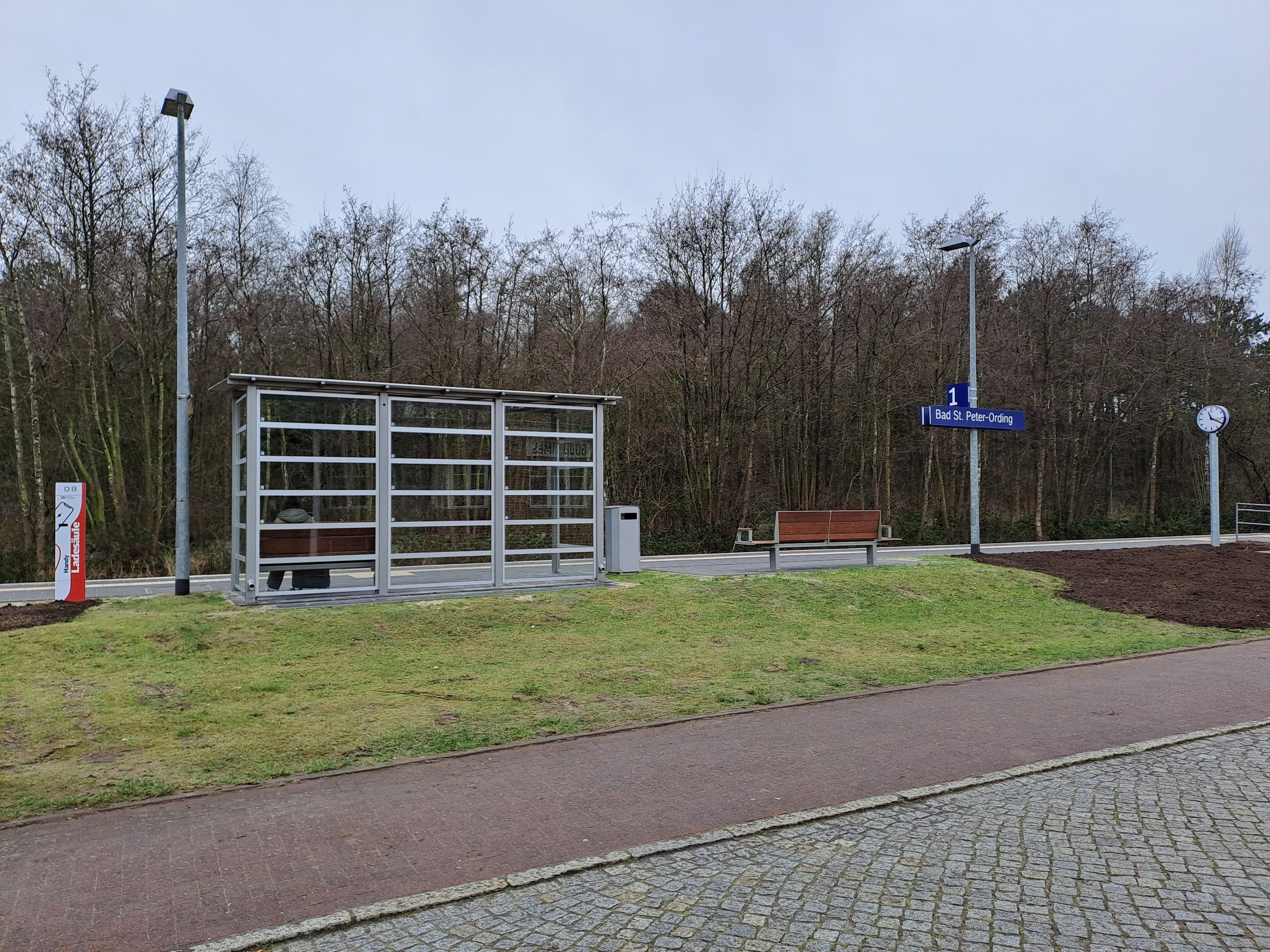 Modernes lichtdurchflutetes Wetterschutzhäuschen und rechts davon eine neue Sitzbank und eine Gleisbeschilderung.