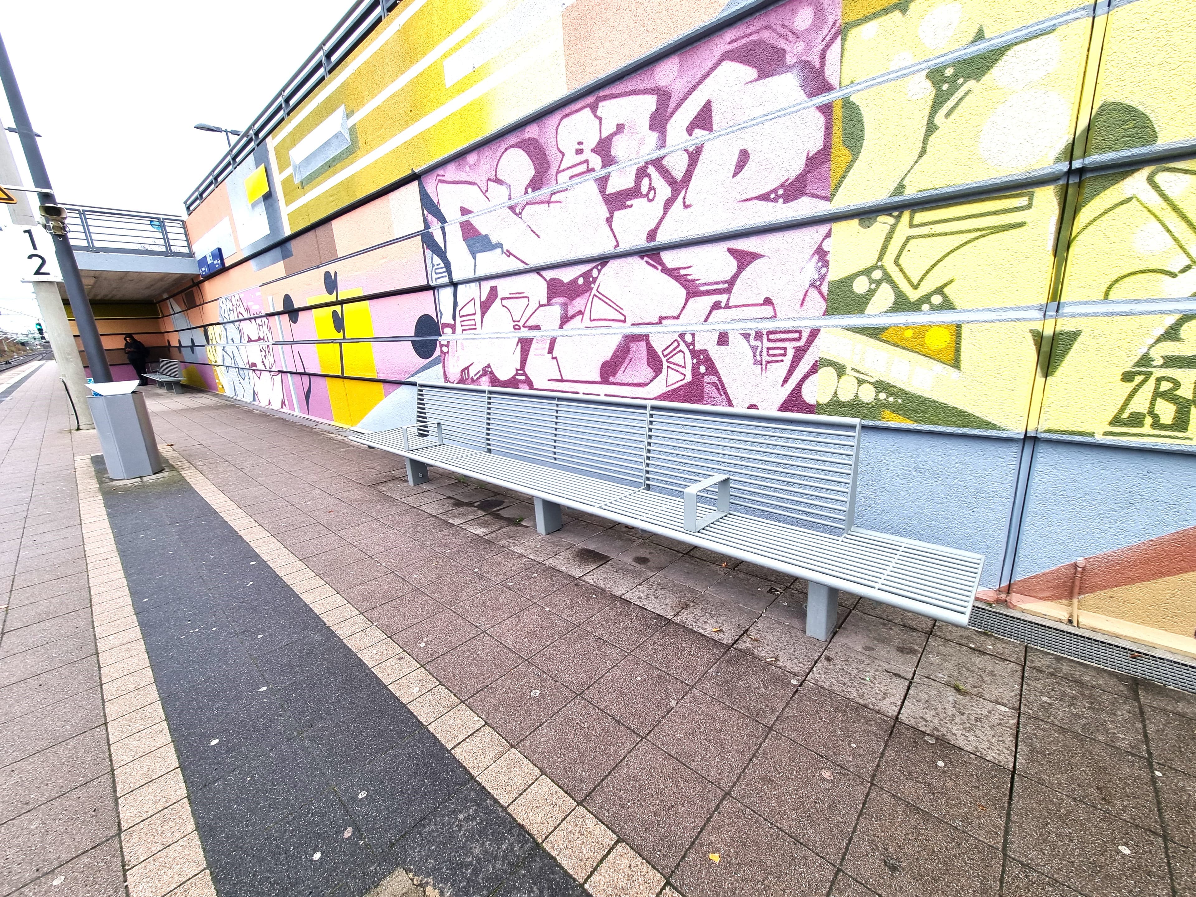 Lange, graue Stahlrohrbank im Vordergrund, im Hintergrund zweite Bank. Beide vor farblich gestalteten Wänden