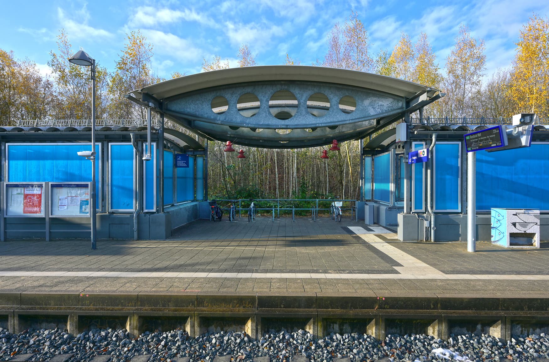 Der Bahnsteig mit dem geschwungenen Bahnhofsdach. Rechts und links davon die neuen, blauen Wände. Auf dem Bahnsteig die Reisendeninformation, eine Vitrine, das Blindenleitsystem und ein mit Fischen bemalter Streugutkasten.