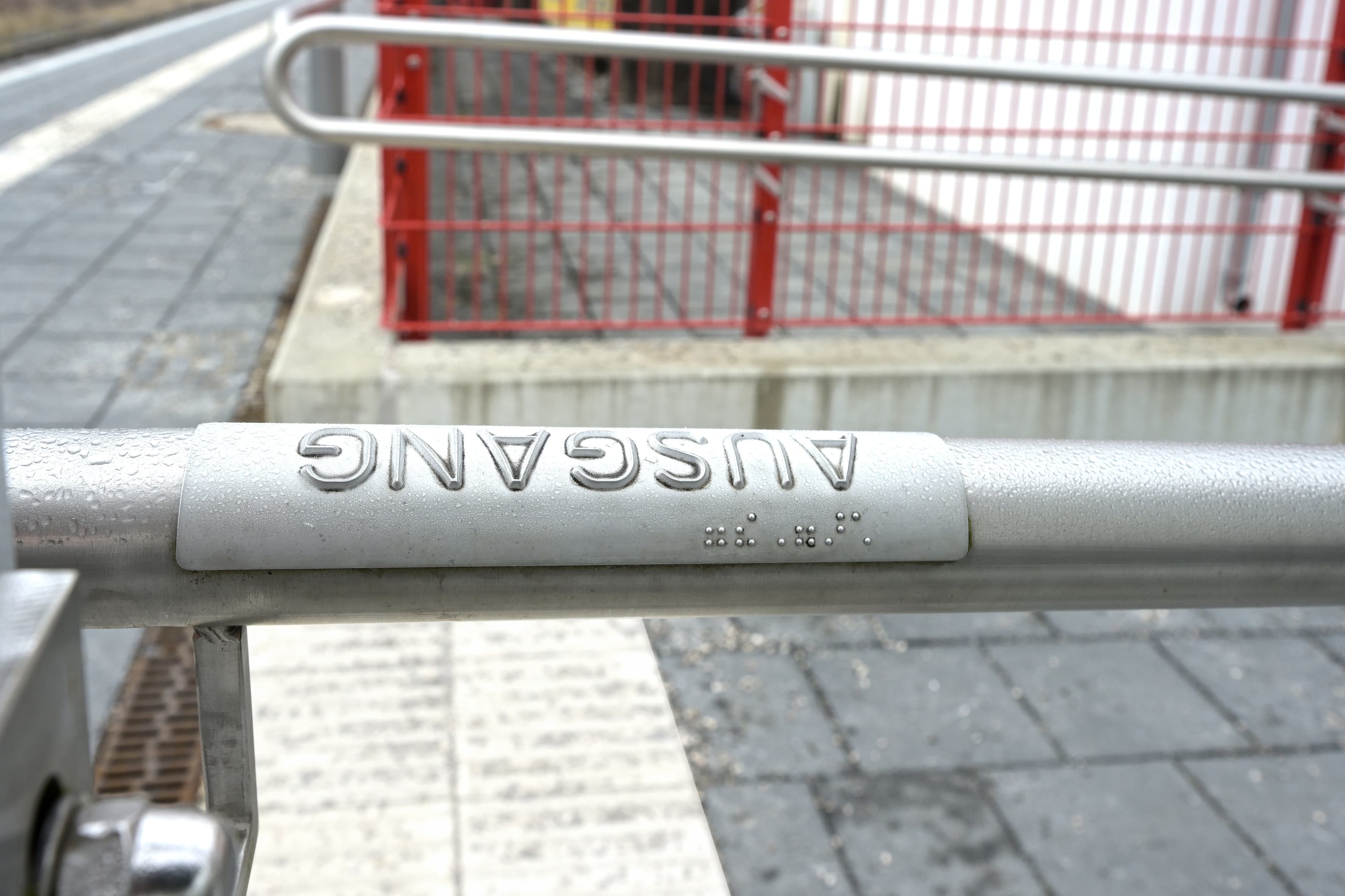 Abgebildet ein Geländer an der Rampe des Zukunftsbahnhofs Hohenpeißenberg. Dieses ist mit einer taktilen Handlaufbeschilderung versehen. Auf dieser ist zum einen Brailleschrift und zum anderen das Wort "Ausgang" in erhabenen Buchstaben.