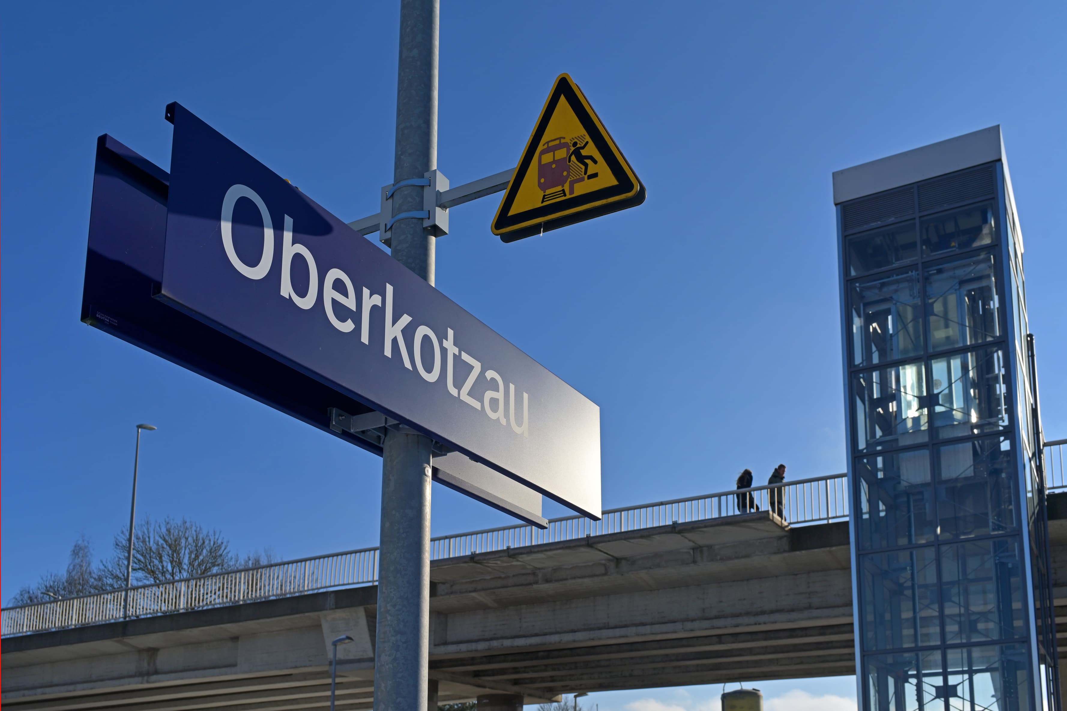 Im Vordergrund das Bahnhofsschild vor wolkenfreiem Himmel. Dahinter ein Aufzugturm, der von den Bahnsteigen auf die angeschlossene Überführung führt. 