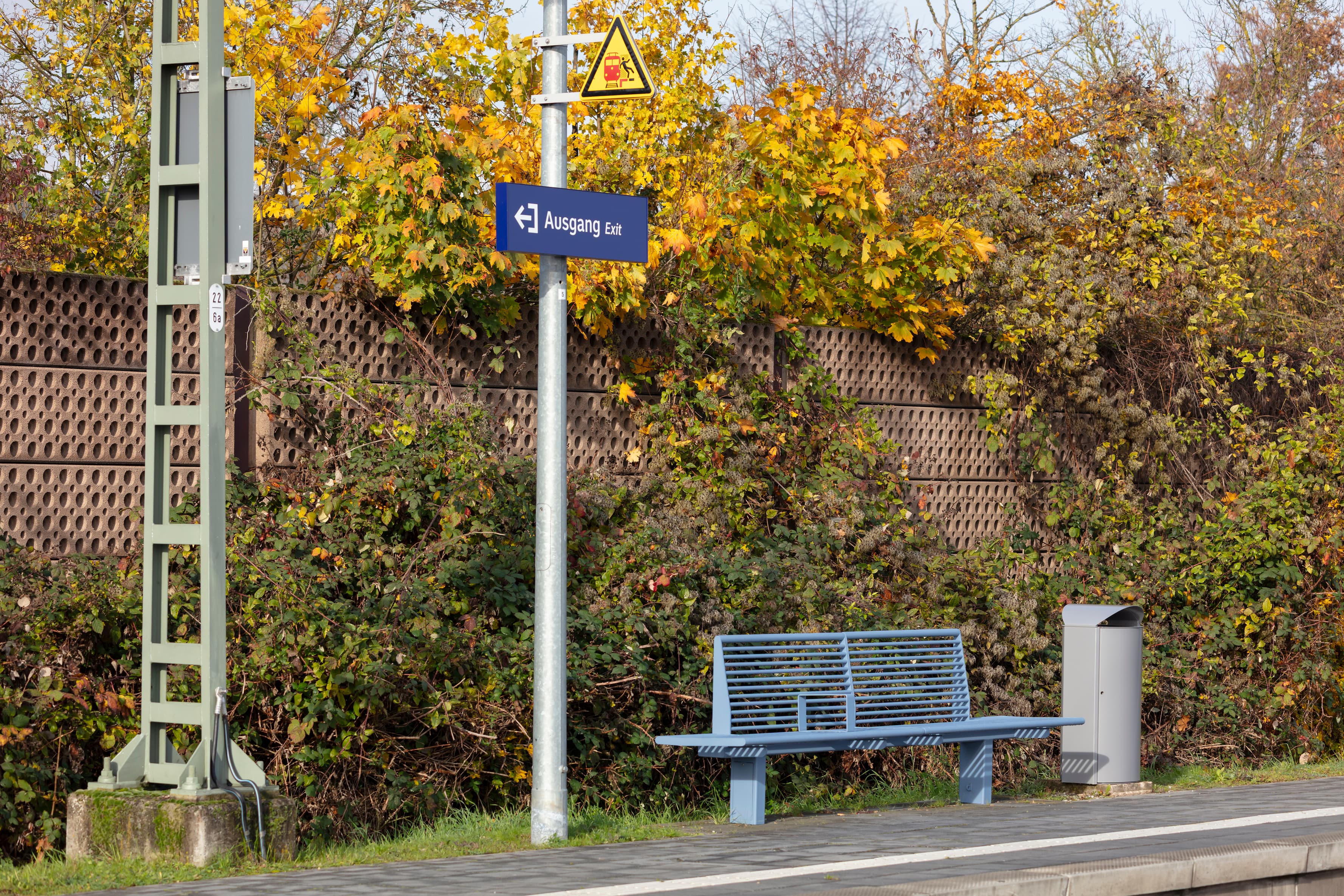 Blaue Sitzbank auf Bahnsteig, daneben dein Mülleimer und eine Stele mit Wegeleitung zum Ausgang.