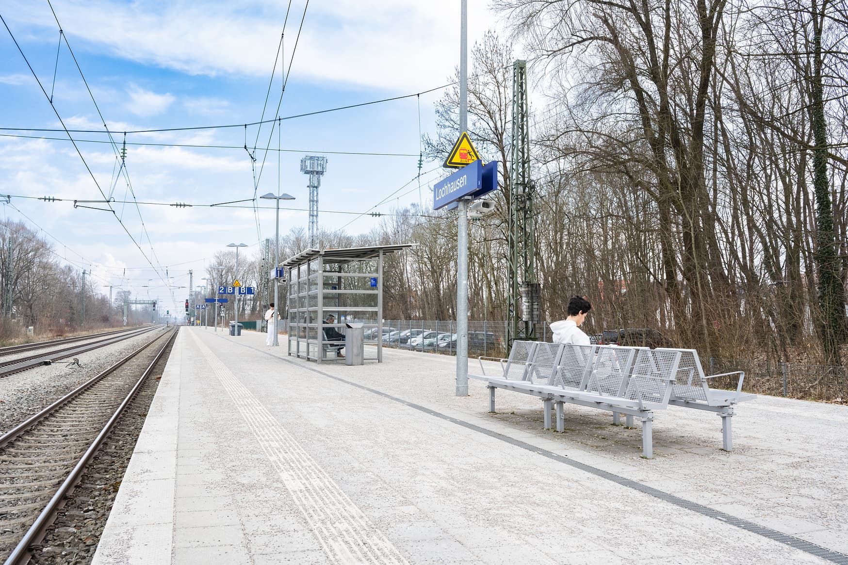 Blick auf einen Bahnsteig. Links davon befindet sich das Gleis. Auf dem Bahnsteig sind mittig Sitzbänke, Masten mit blauen Schildern und Wetterschutzhäuschen zu sehen.