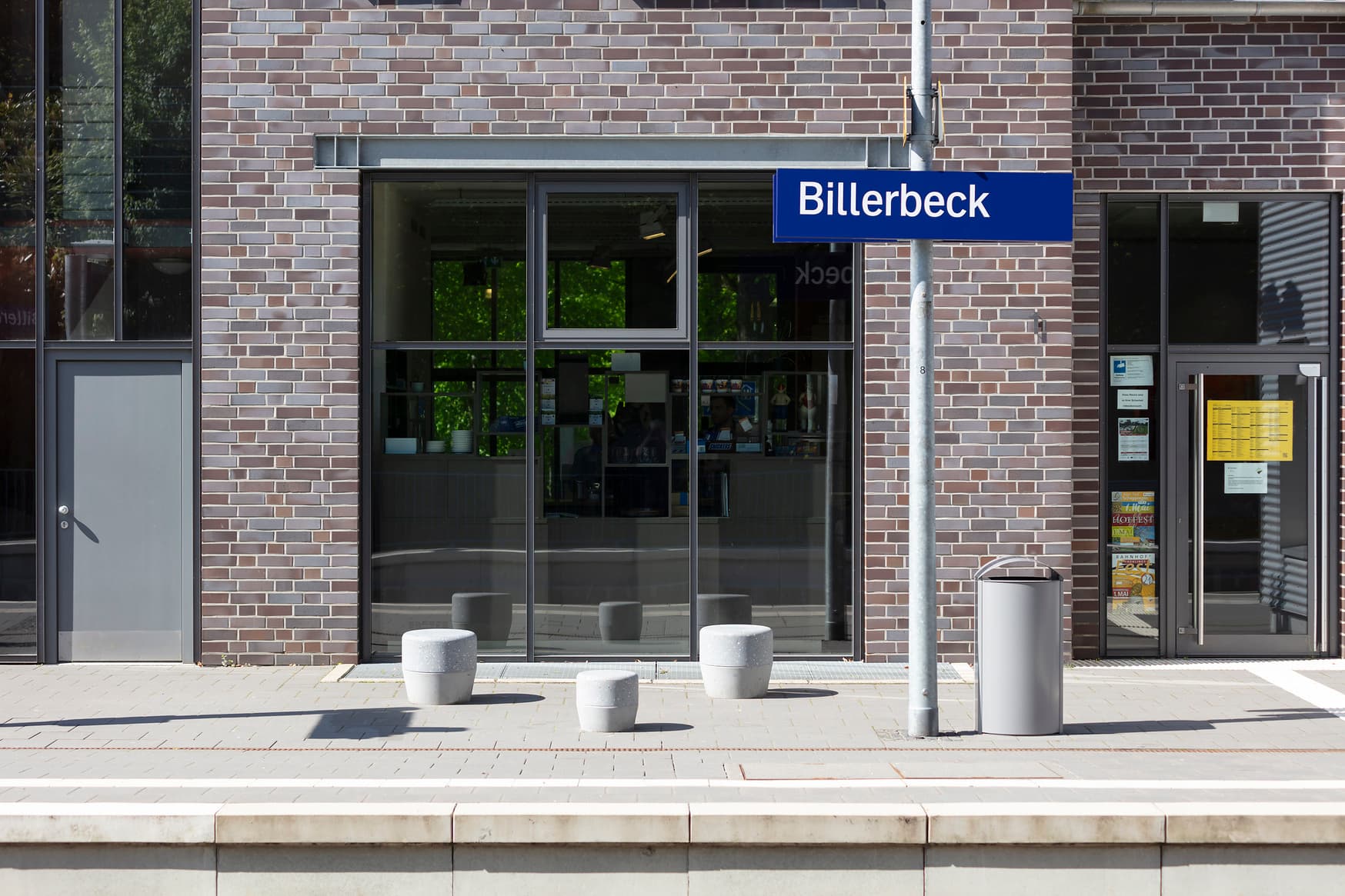Drei Betonhocker auf dem Bahnsteig im Dreieck angeordnet vor dem Empfangsgebäude und neben dem Bahnhogsschild.