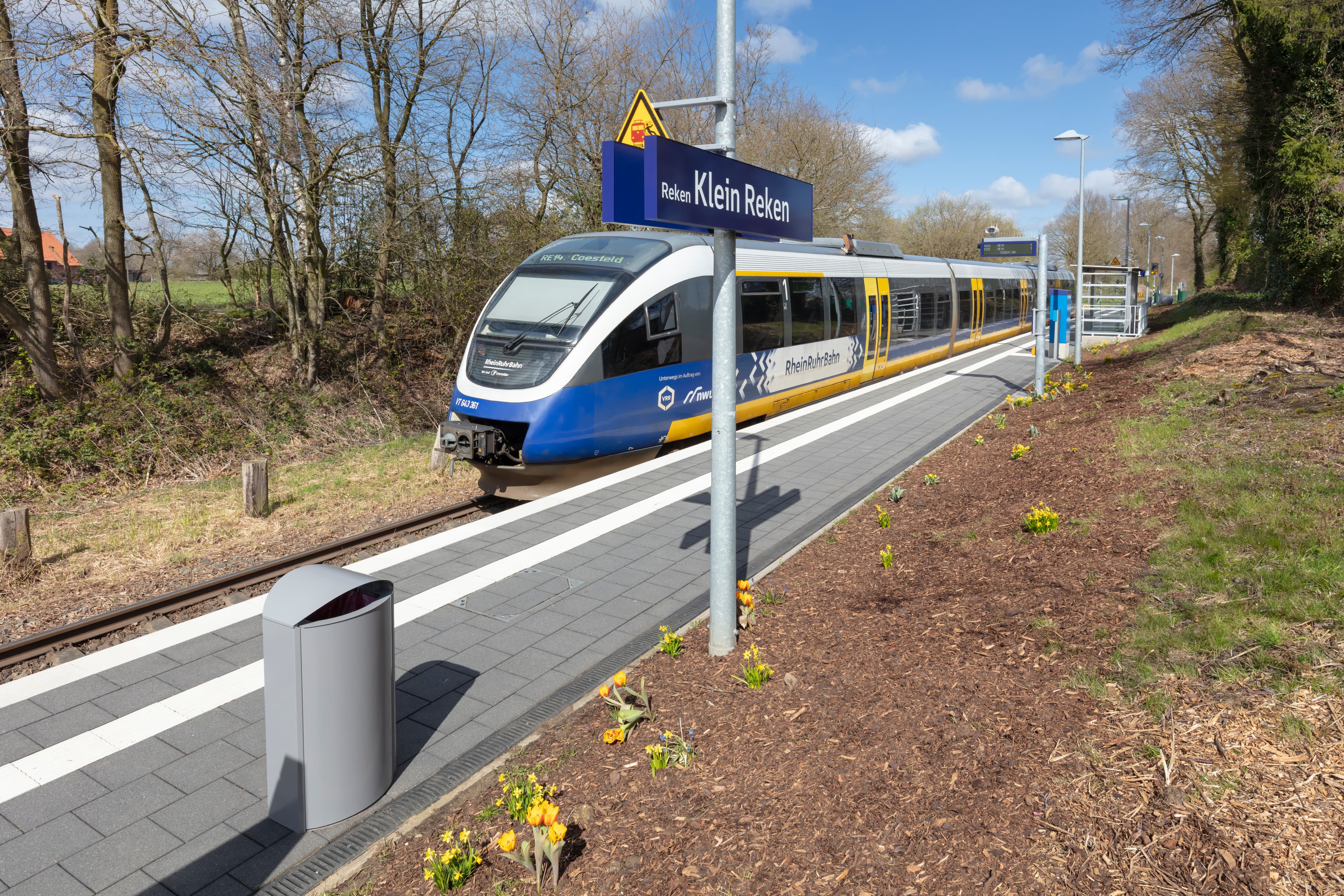 Grünfläche mit Blumen neben einem Bahnsteig. Dahinter in blauer Zug. 