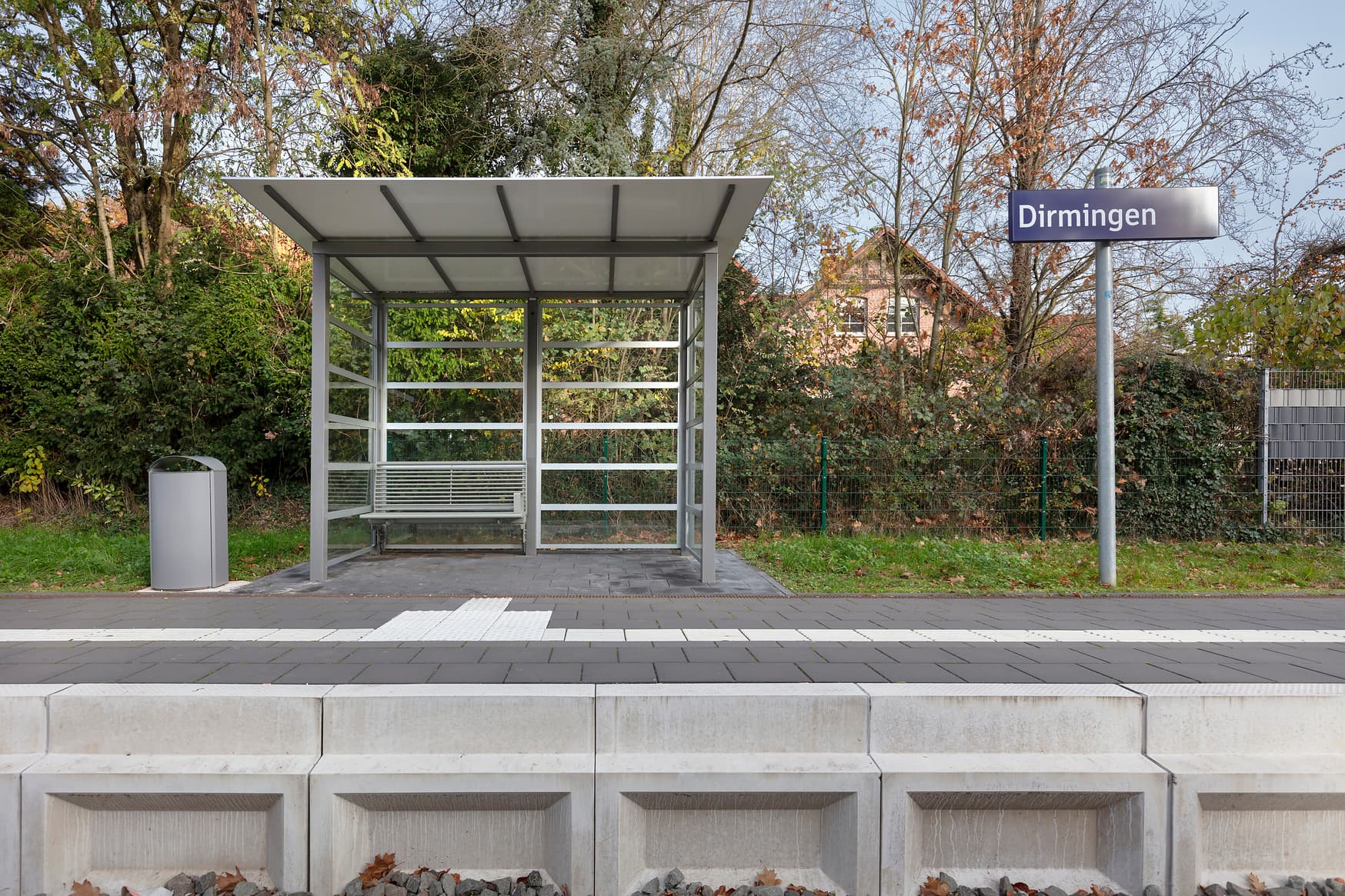 Das volltransparente Wetterschutzhaus auf dem Bahnsteig, rechts daneben die Stele mit dem Bahnhofsschild Dirmingen, links daneben ein Mülleimer.
