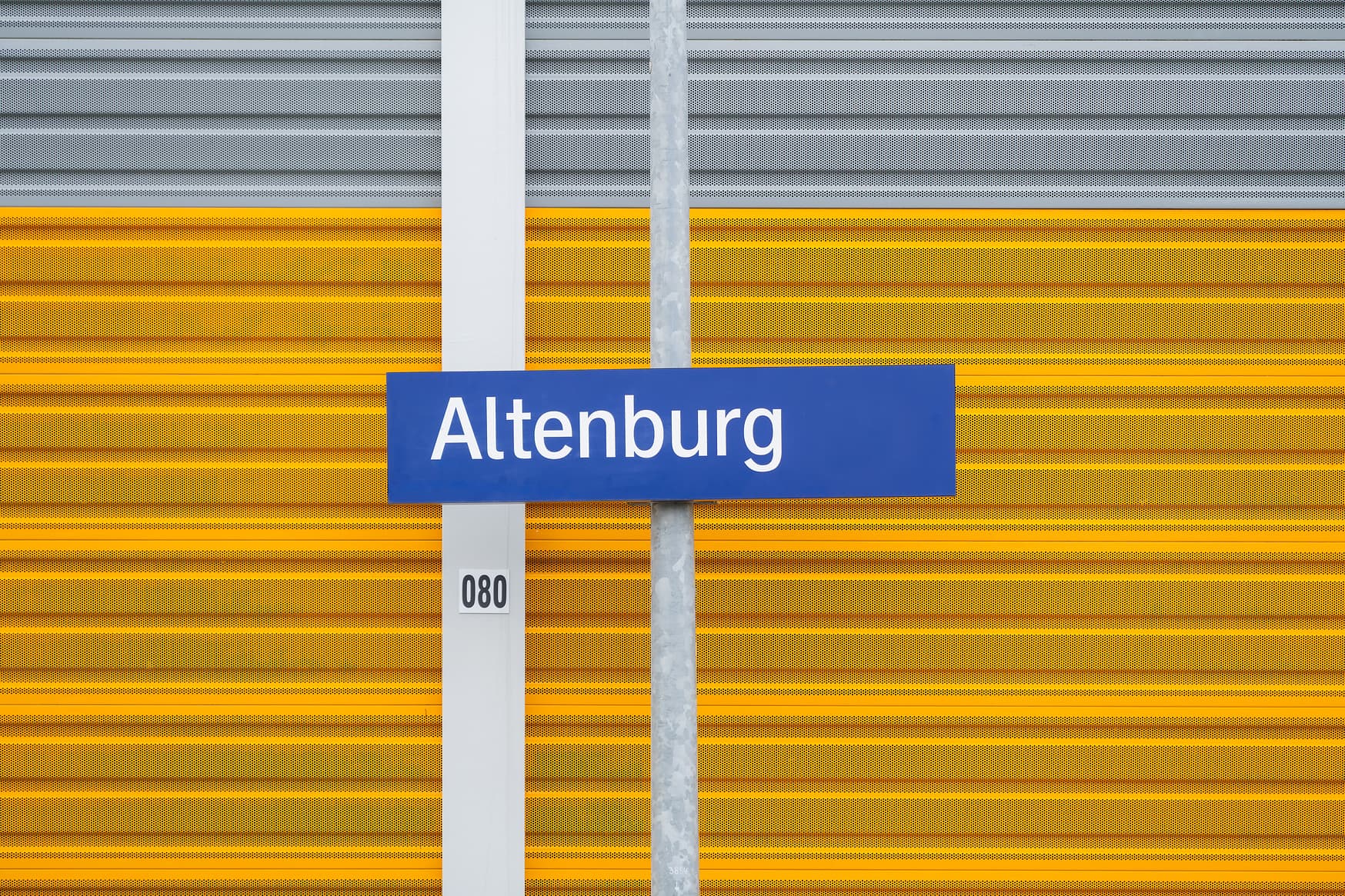 Das Schild mit dem Bahnhofsnamen des Bahnhofs Altenburg vor einer gelben Lärmschutzwand.