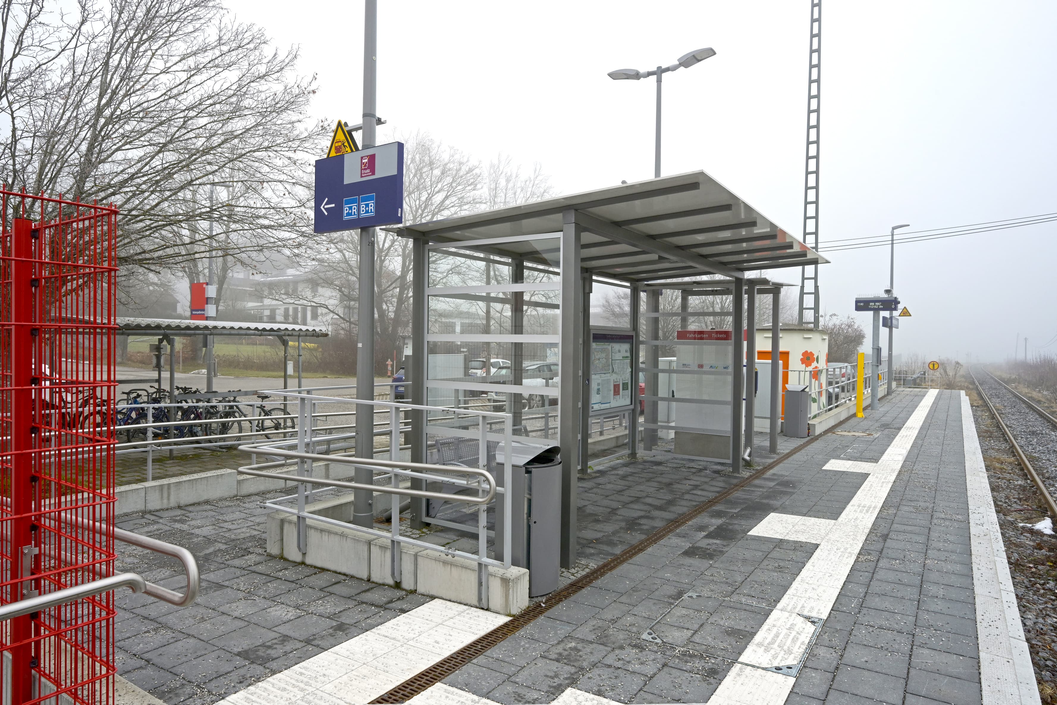  Abgebildet der Bahnsteig des Zukunftsbahnhofs Hohenpeißenberg. Im Zentrum des Bildes ist das Wetterschutzhaus mit transparenten Wänden. Auf der rechten Seite der Bahnsteig. Links im Bild eine Rampe, die zum Bahnsteig führt.