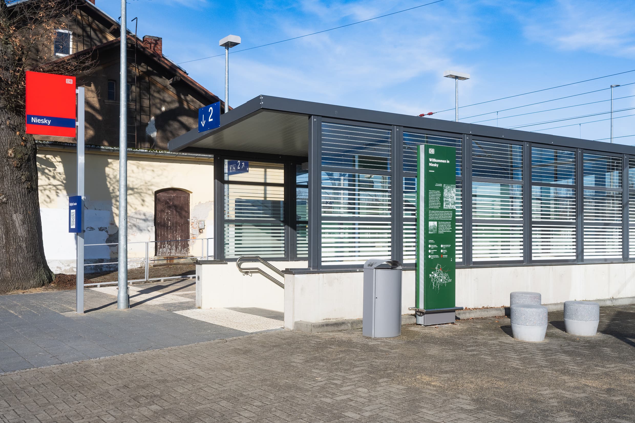 Blick auf den Zugang zum Tunnel. Neben der Treppeneinhausung steht ein Mülleimer, drei Betonhocker und eine grüne Stele mit Infos zur Region
