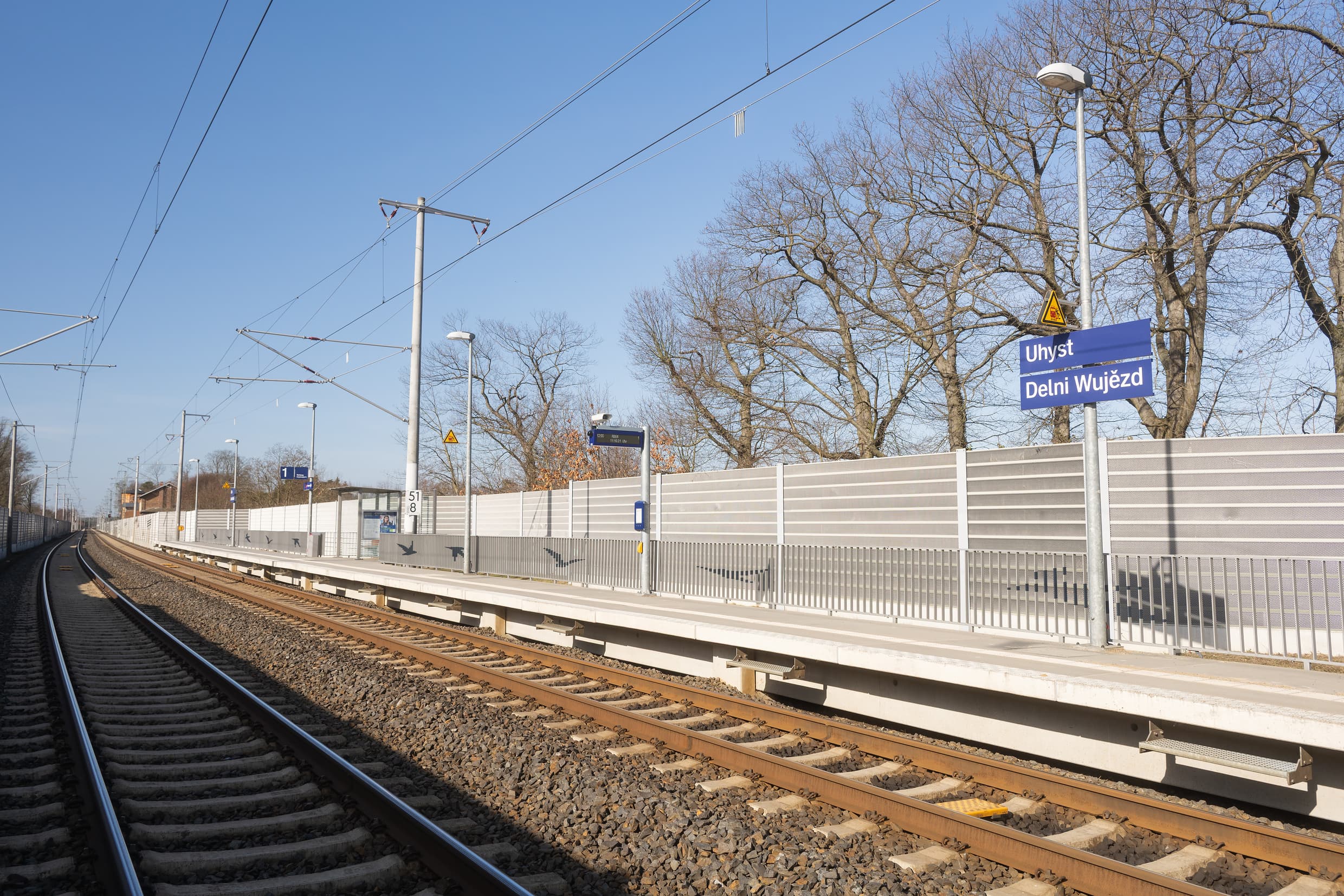 Blick auf die gesamte Verkehrsstation: Die Vogelsilhouetten auf den Geländern sind gut zu sehen. Gleichzeitig auch das Bahnhofsnamensschild, der digitale Anzeiger und weiter hinten das Wetterschutzhaus.