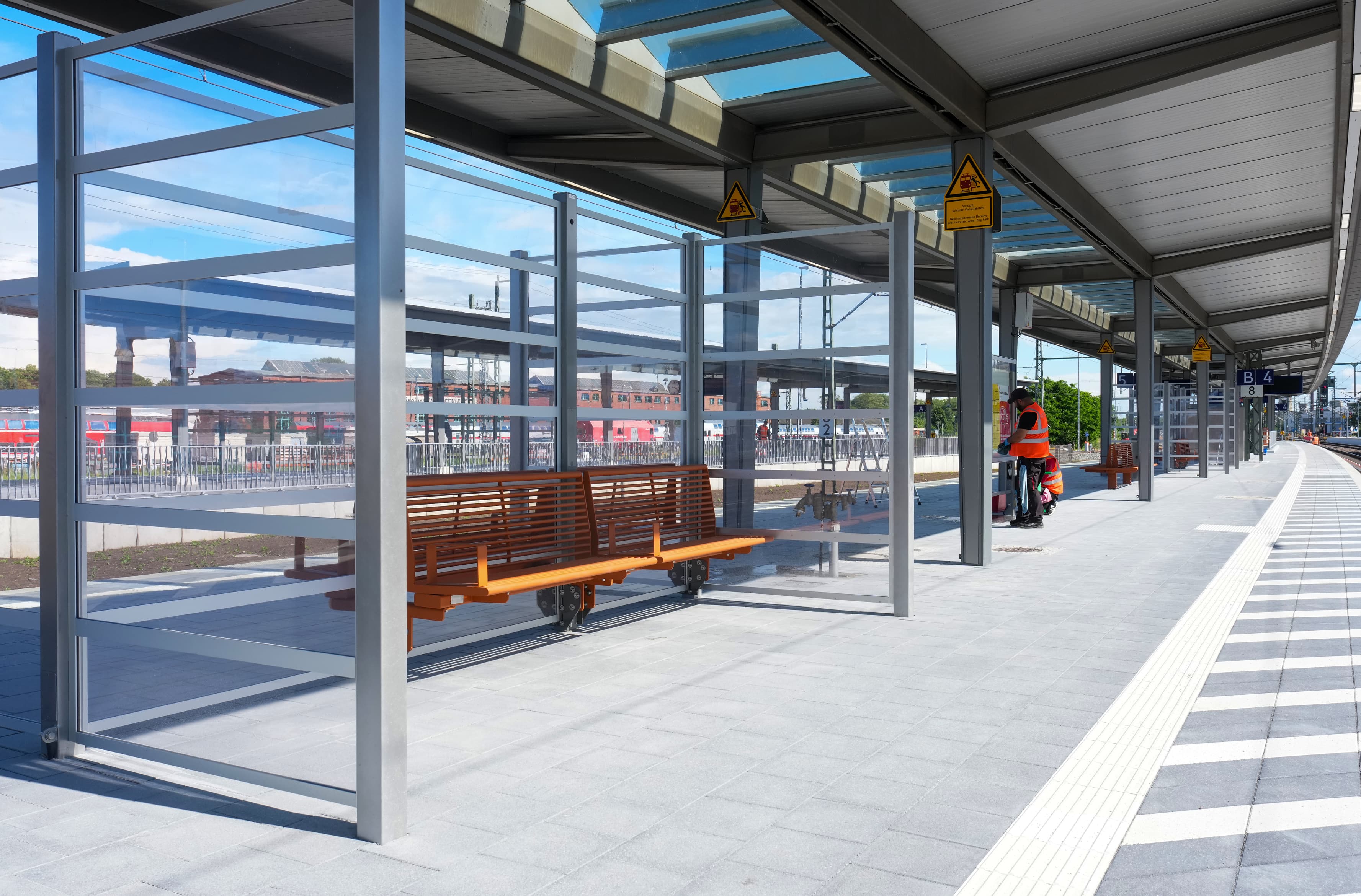 Ein Wartehaus aus Glas mit einer orangenen Sitzbank an einem Bahnsteig.