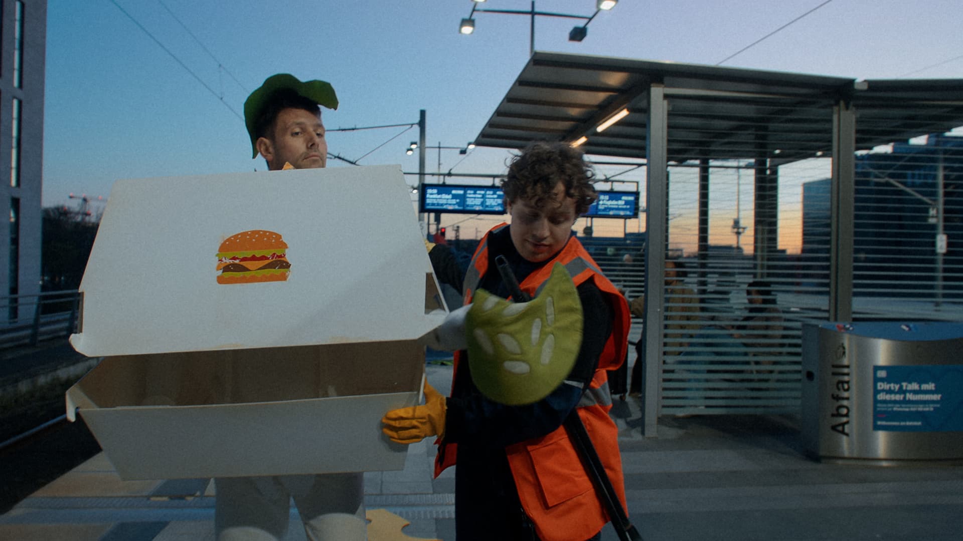 A person dressed as a burger is taken away by a cleaner and demonstratively throws a cucumber on the floor while walking.