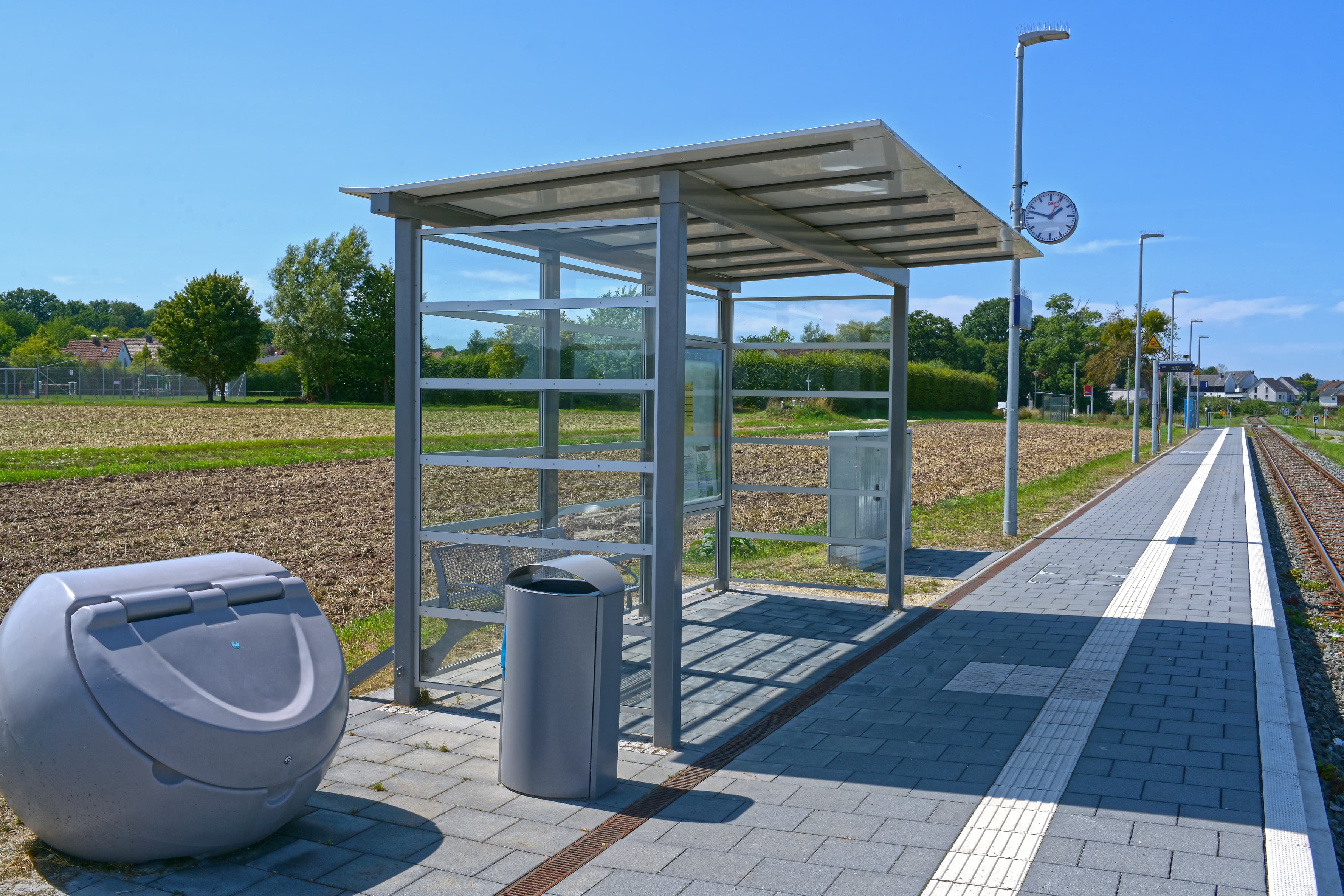 Zu sehen ist ein Unterstand aus Glas mit einer Bank aus Metall im Inneren. Links davor steht ein Streugutbehälter und ein Mülleimer. Im Hintergrund ein ein Beleuchtungsmast mit der Bahnhofsuhr zu sehen. Rechts im Bild verlaufen die Gleise entlang des Bahnsteigs.