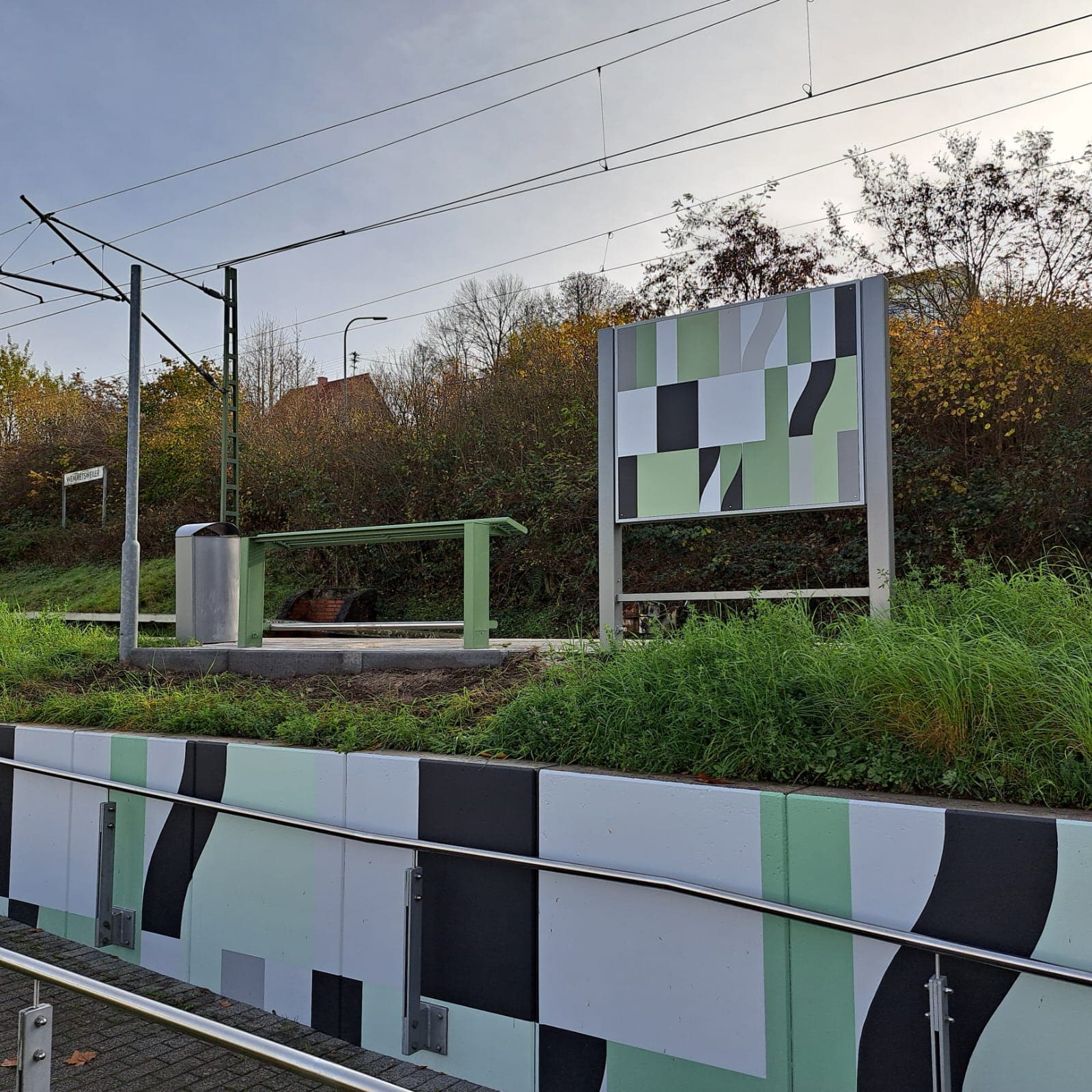 Blick auf bunt gestaltete Stützmauer mit Handlauf. Dahinter befinden sich oberhalb ein grüner Anlehner und eine bunt gestaltete Vitrinenrükseite.
