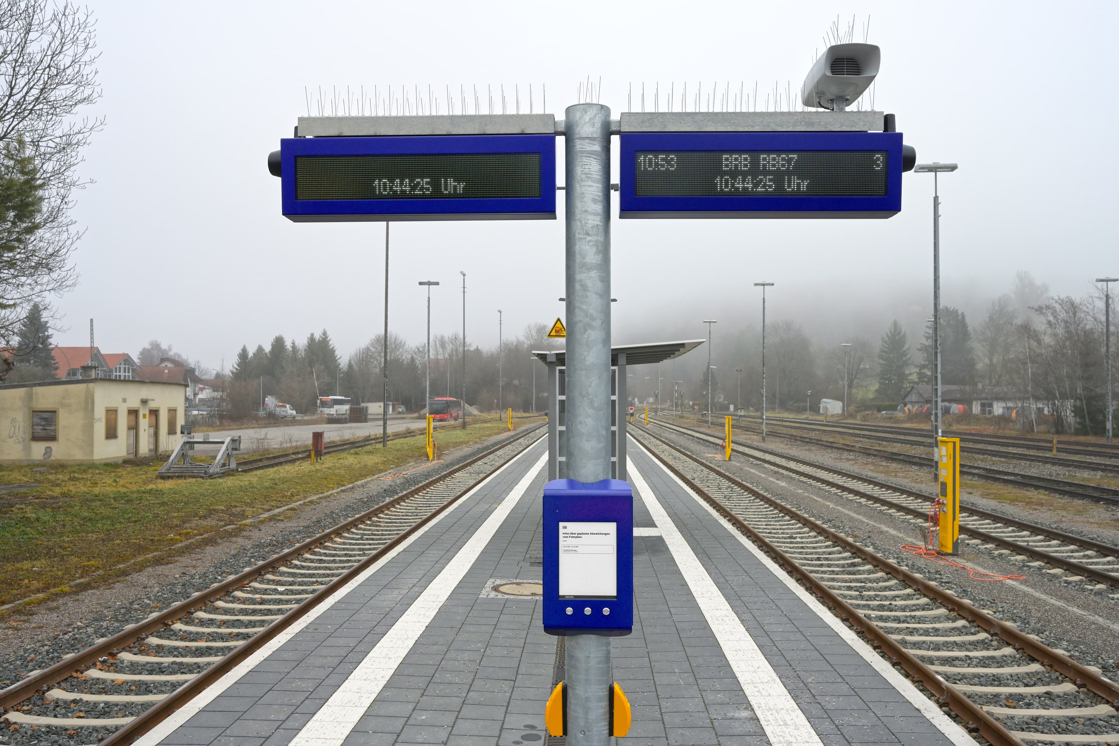 Abgebildet ein Bahnsteig. In der Mitte des Bildes ein Mast an dem digitale Anzeigegeräte zur Fahrgastinformation befestigt sind. Rechts und links jeweils Gleise. Im Hintergrund ein Wetterschutzhaus.