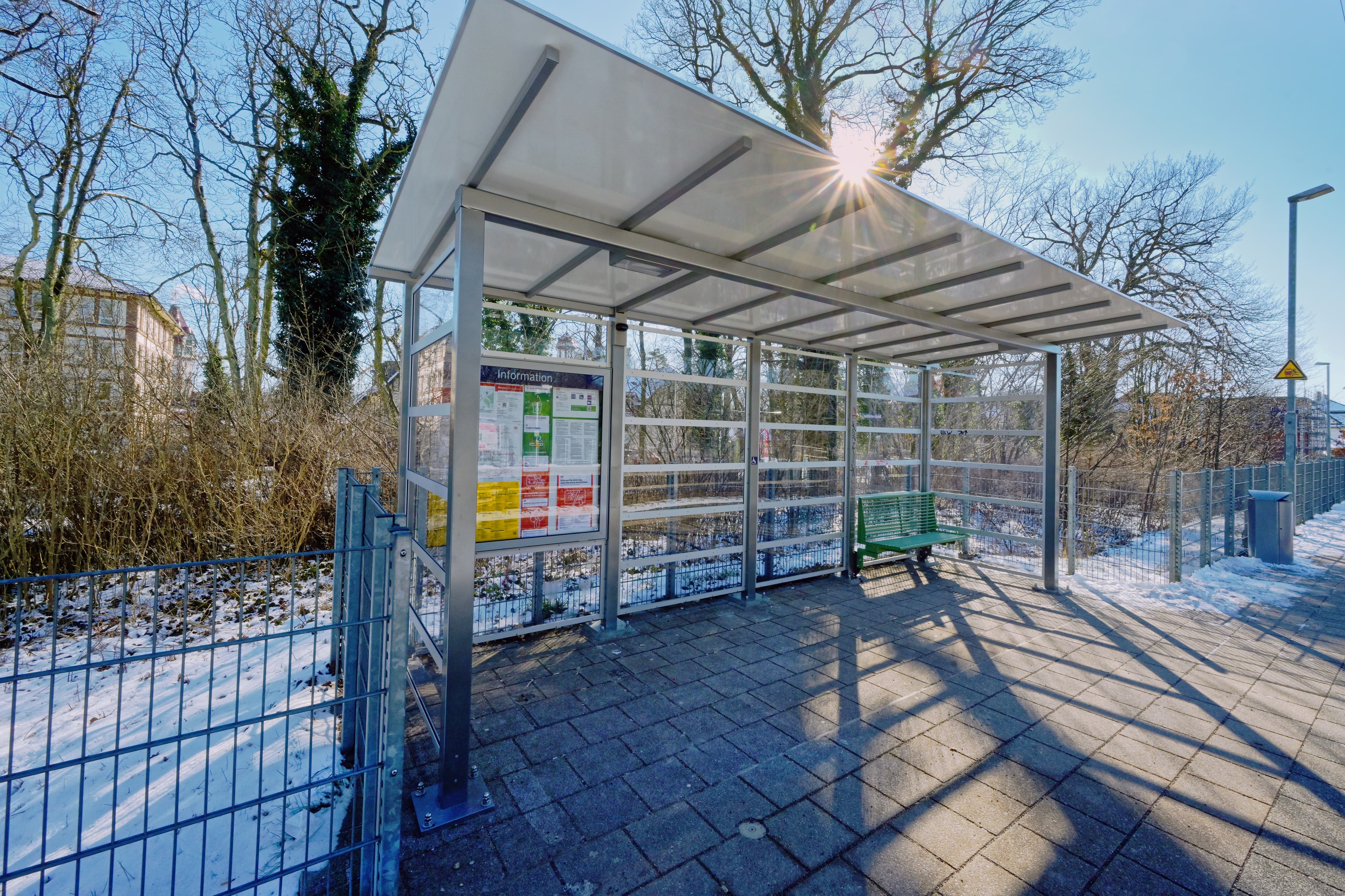 Transparentes Wetterschutzhäuschen mit grüner Bank und Info-Kasten.