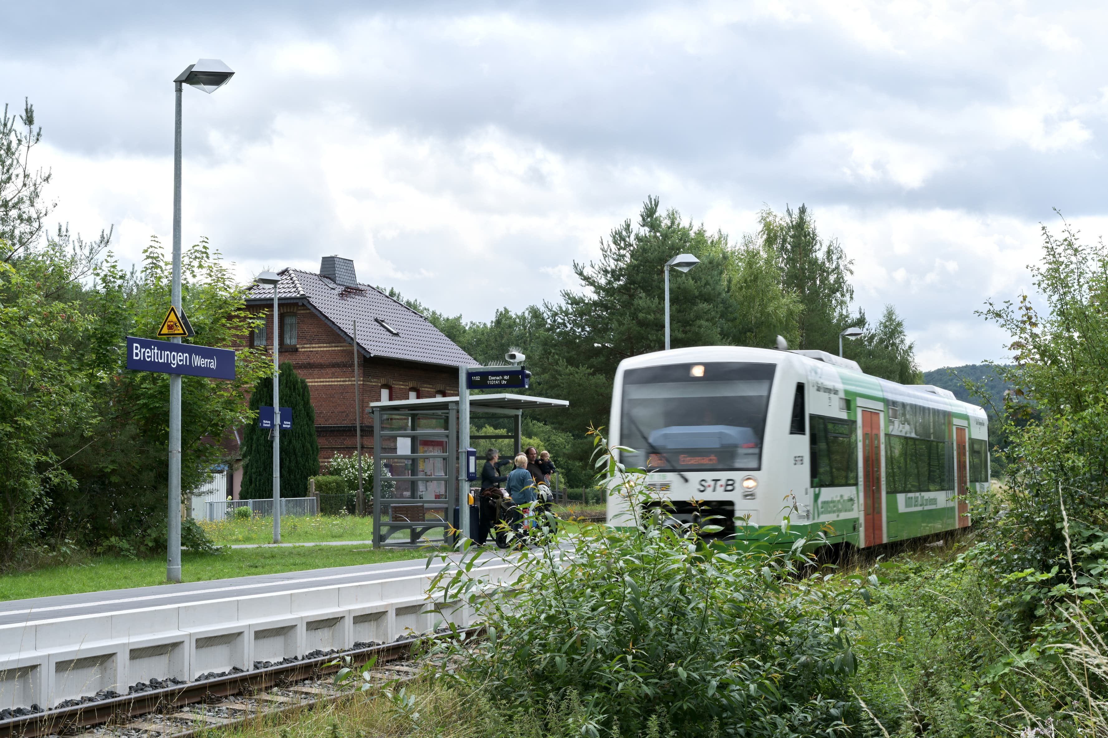 Einahrender Zug an neu gebautem Bahnsteig. Reisende warten am voll transparenten und neuem Wetterschutzhaus auf den Einstieg.