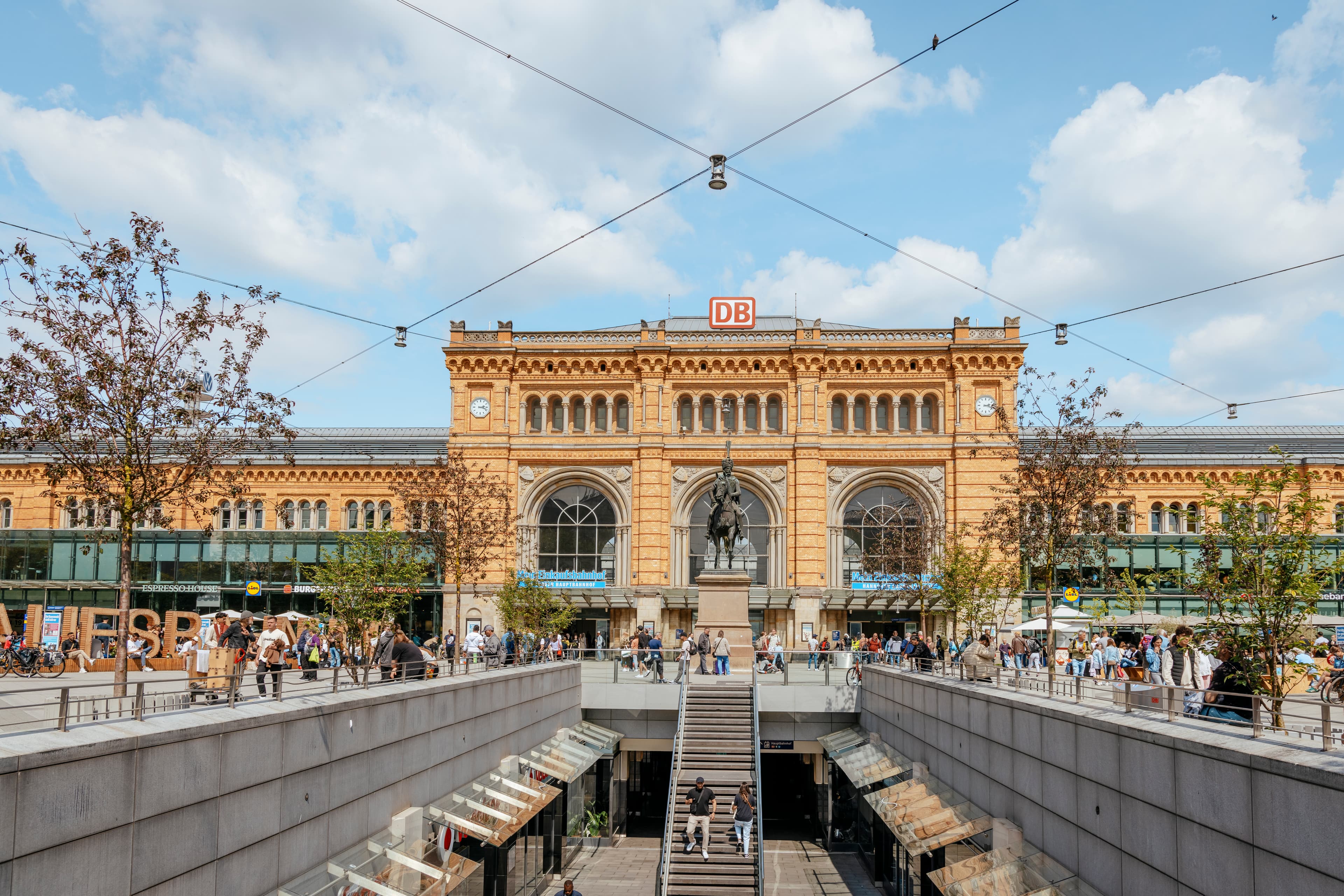 Blick auf das historische Bahnhofsgebäude des Hannover Hbf und das Pferde-Denkmal von Ernst-August. Rechts und links entlang des Passarellenabgang stehen kleine Bäume auf dem Bahnhofsvorplatz. Es sind einige Menschen bei blauem leicht bewölktem Himmel unterwegs.