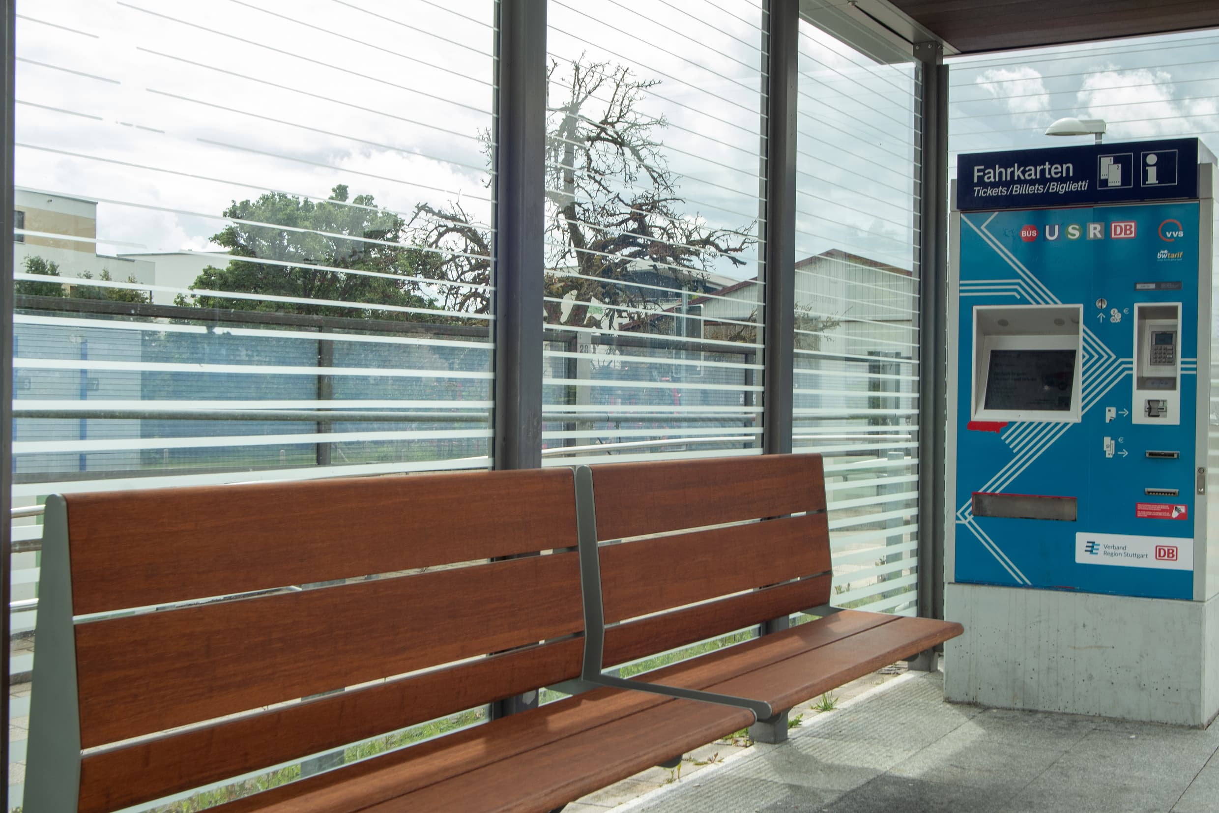 Wetterschutzhaus mit Glaswänden, Sitzbank aus Holz und daneben ein Fahrkartenautomat.
