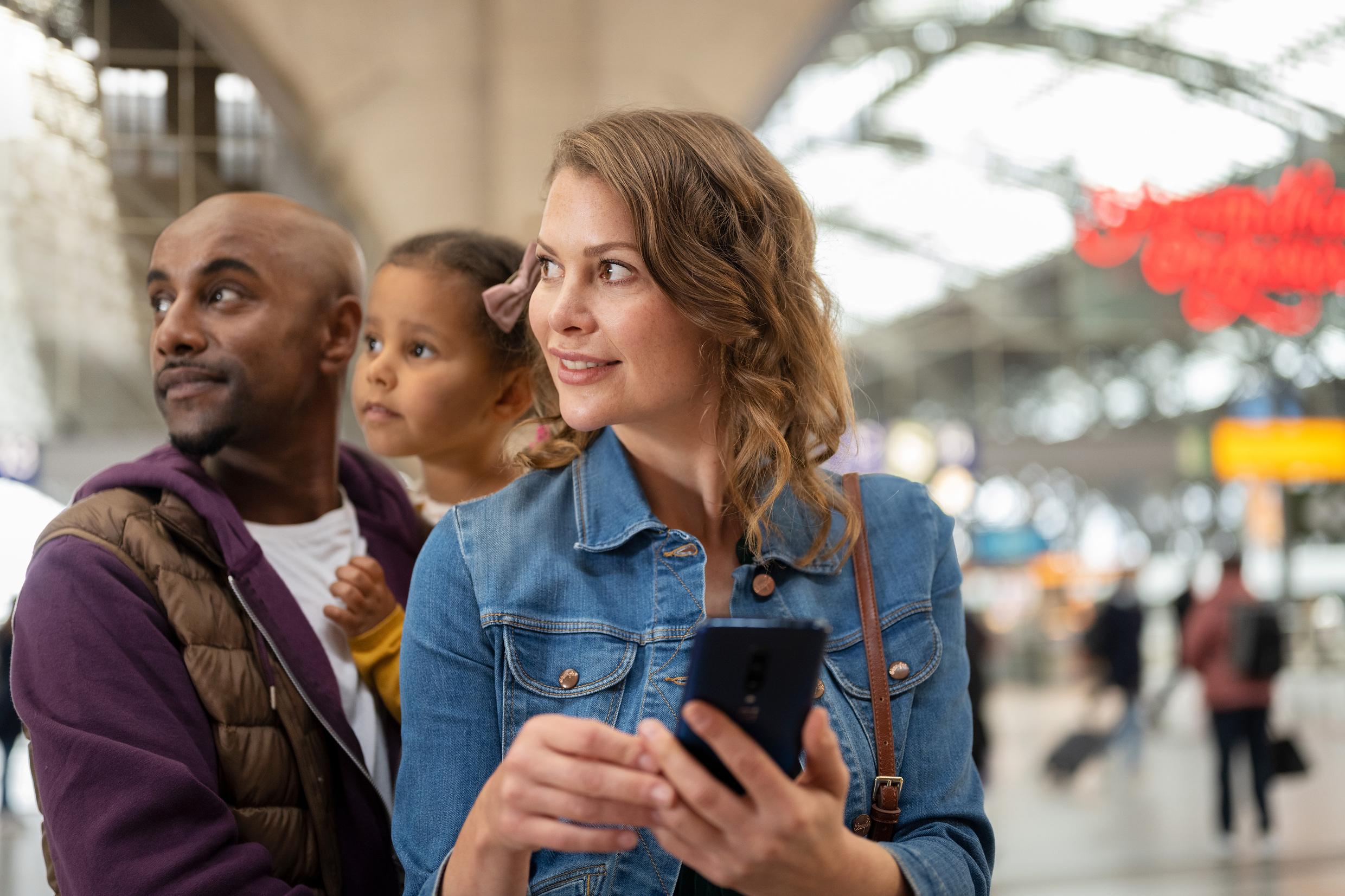 Ein Mann mit einem Mädchen auf dem Arm und eine Frau mit einem Smartphone am Bahnhof.