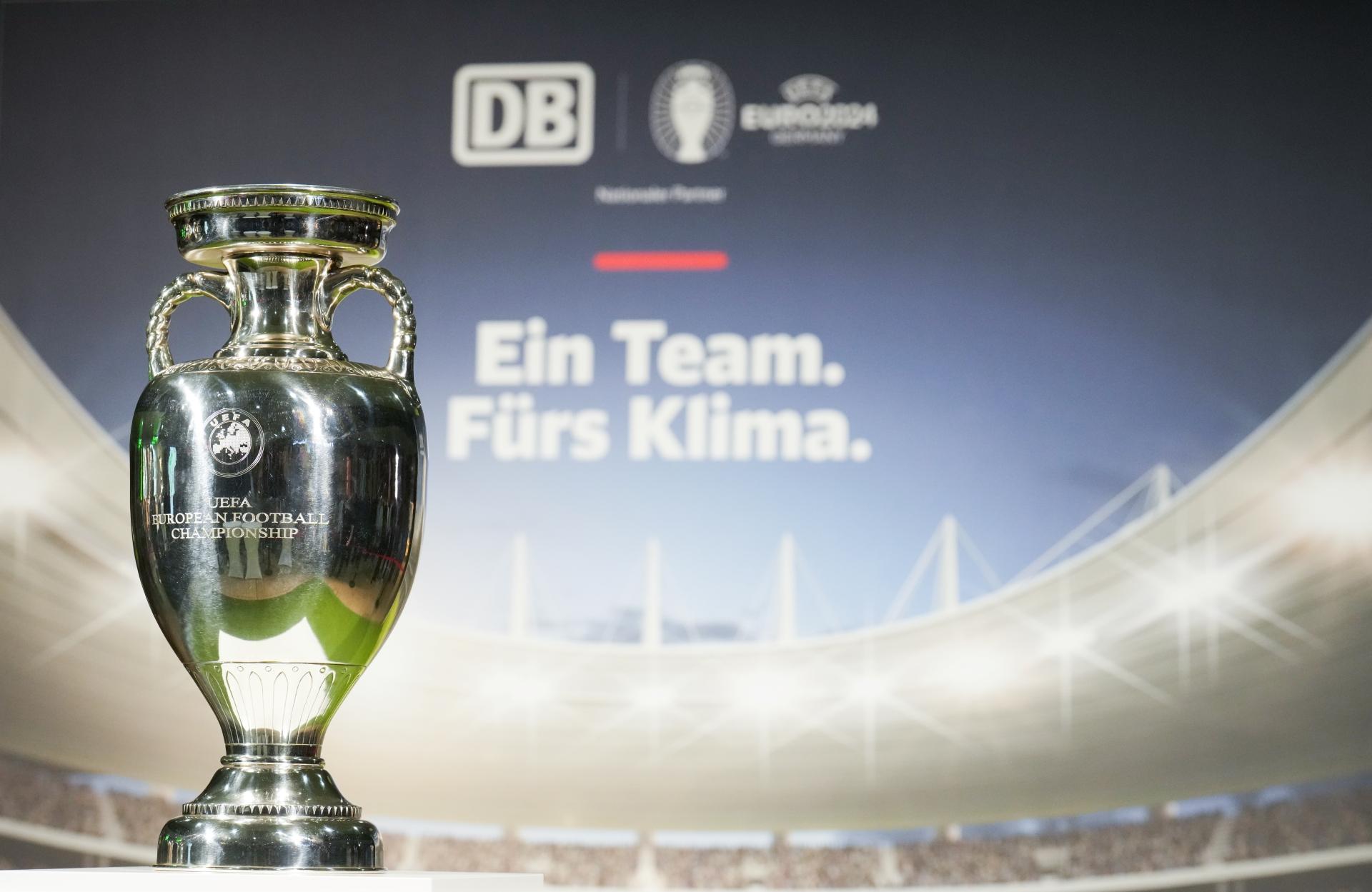 Der EM-Pokal steht auf einem Podest, im Hintergrund steht der Schriftzug "Ein Team. Fürs Klima" mit dem DB Logo und dem Logo der UEFA Euro 2024.	