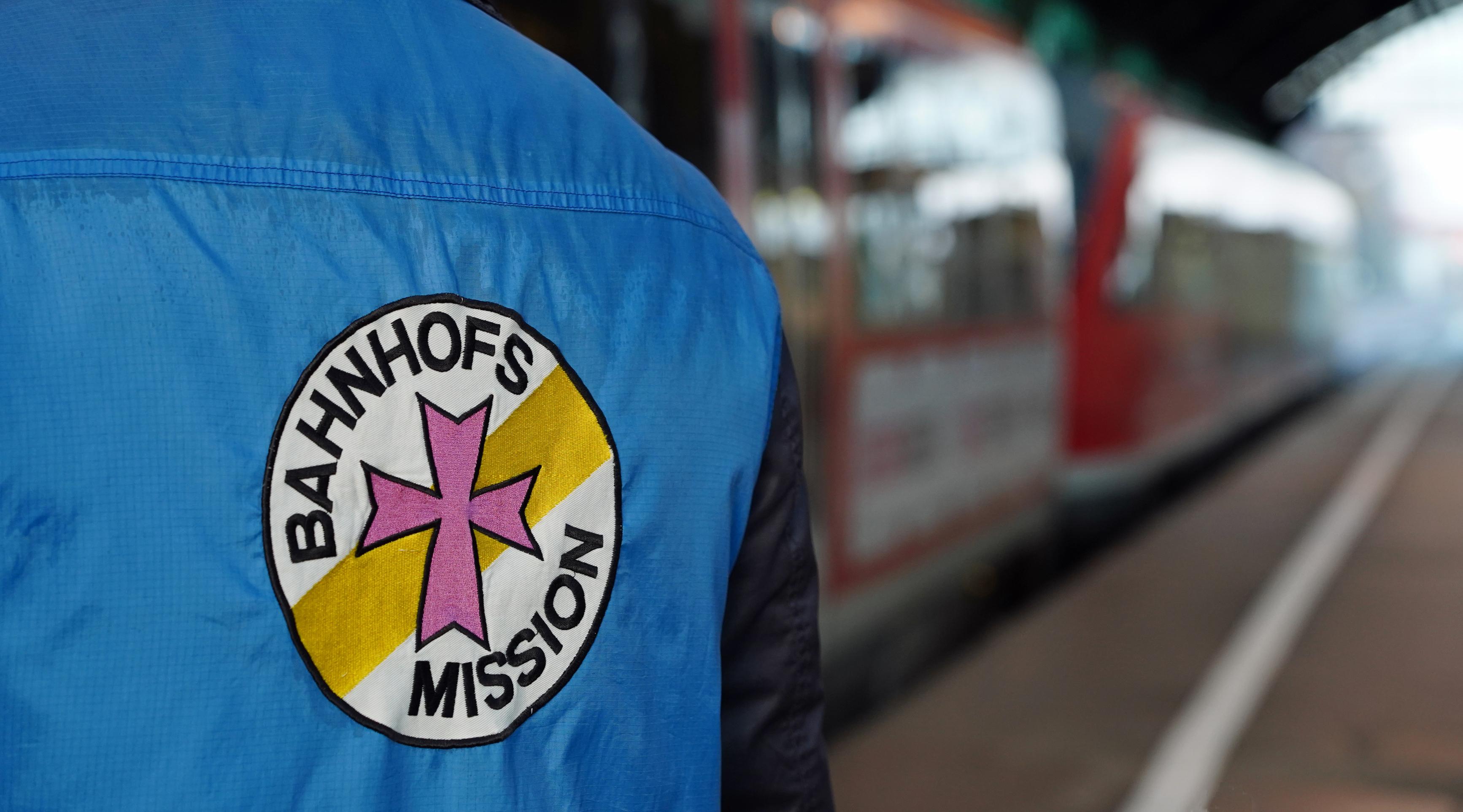 A person on the platform wears a vest with the logo of Bahnhofsmission on the back.