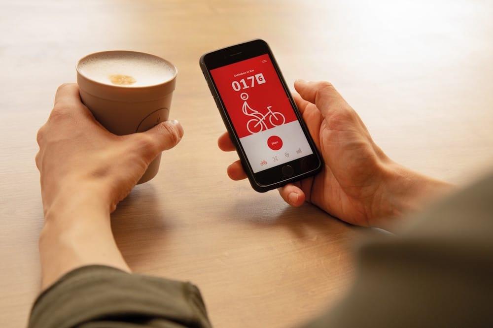 Ein Mann hält in der linken Hand einen Kaffeebecher und in der rechten Hand ein Smartphone, auf dem die App DB Rad+ geöffnet ist.
